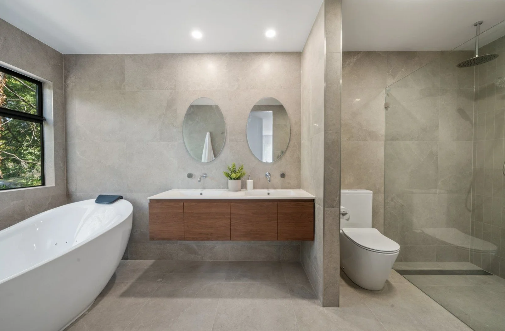Modern bathroom with large window, freestanding bathtub, double vanity with oval mirrors, potted plant, soap dispenser, toilet, and glass walk-in shower with rain showerhead.