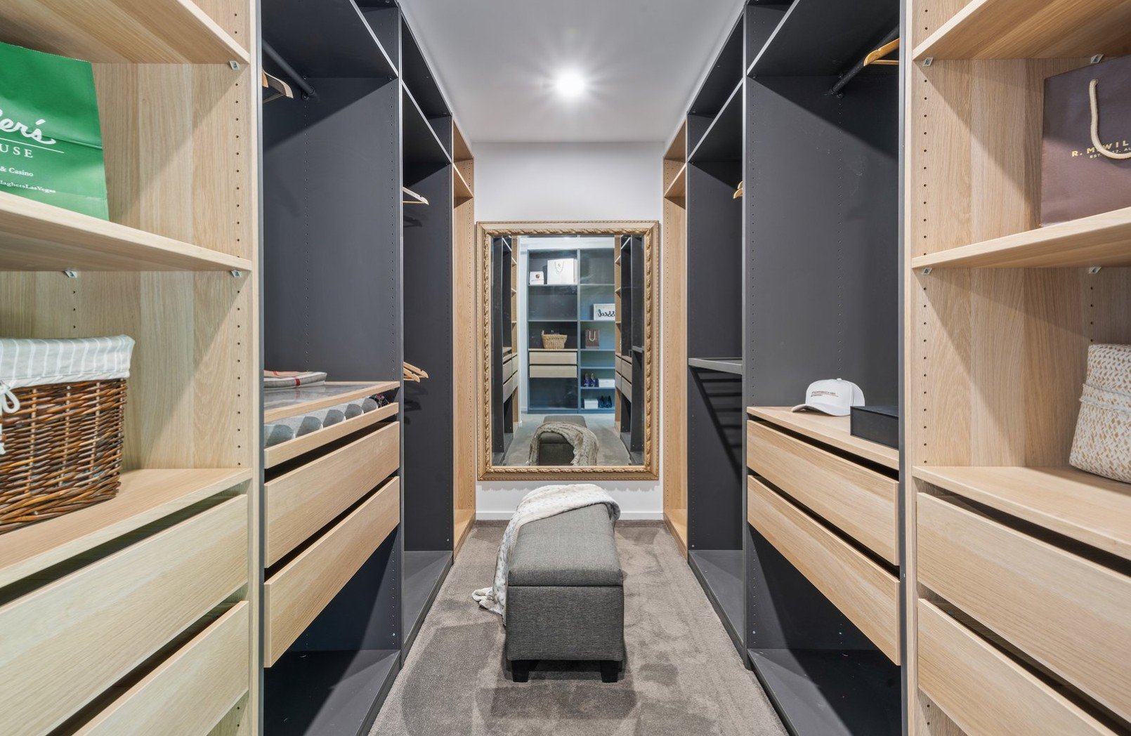 A walk-in closet with wooden and black shelving on both sides, a large ornate mirror at the end, a cushioned bench in front of the mirror, and various personal items like handbags, a hat, and clothing.
