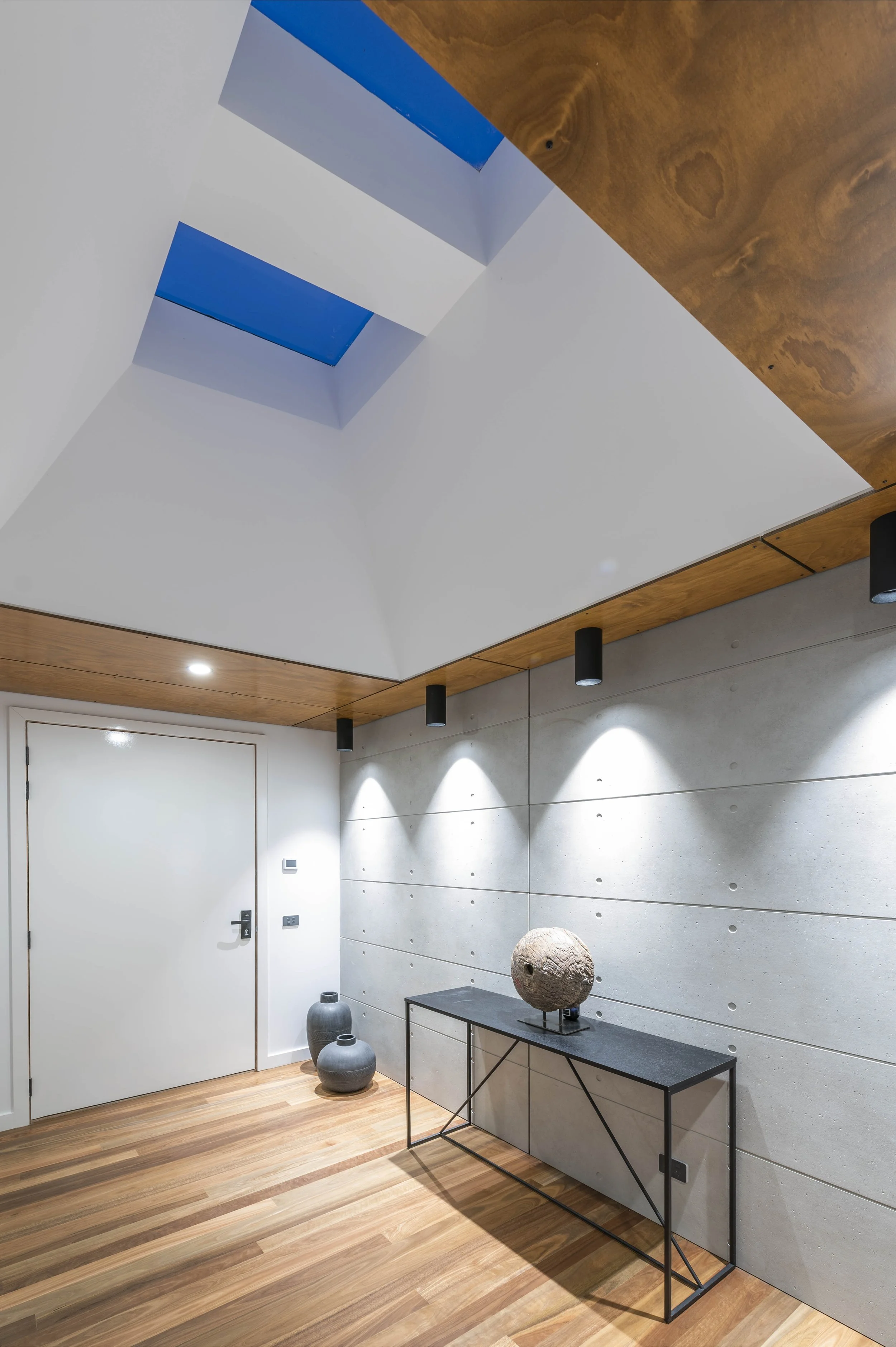 Modern interior room with wooden flooring, a white door, decorative vases on the floor, a black metal console table with a stone sculpture, a textured concrete wall, a wooden ceiling with recessed lighting, and a ceiling skylight.