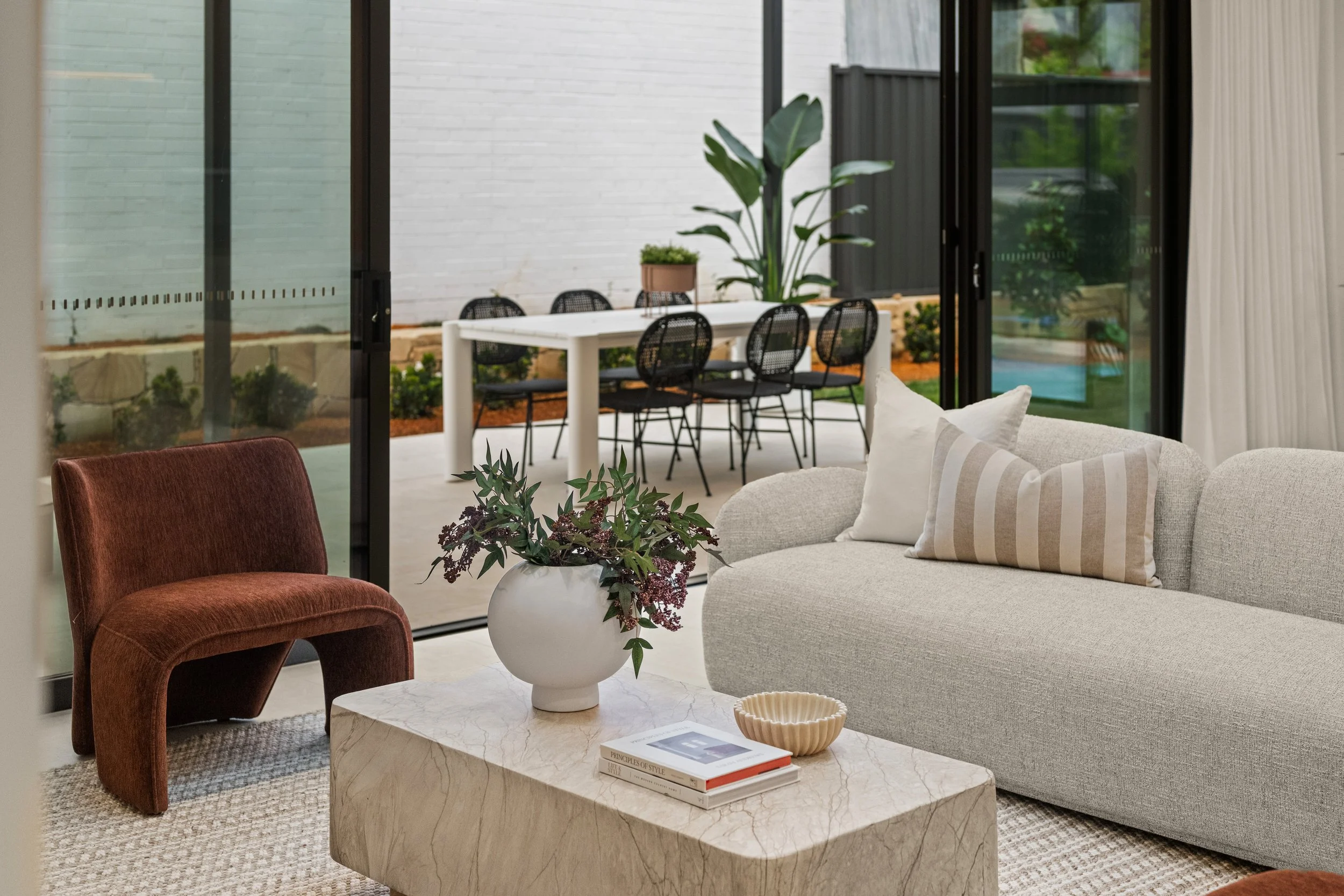 Living room with a beige sofa, a red velvet armchair, and a marble coffee table with books and a plant, overlooking an outdoor dining area with a white table and black chairs through large glass sliding doors.