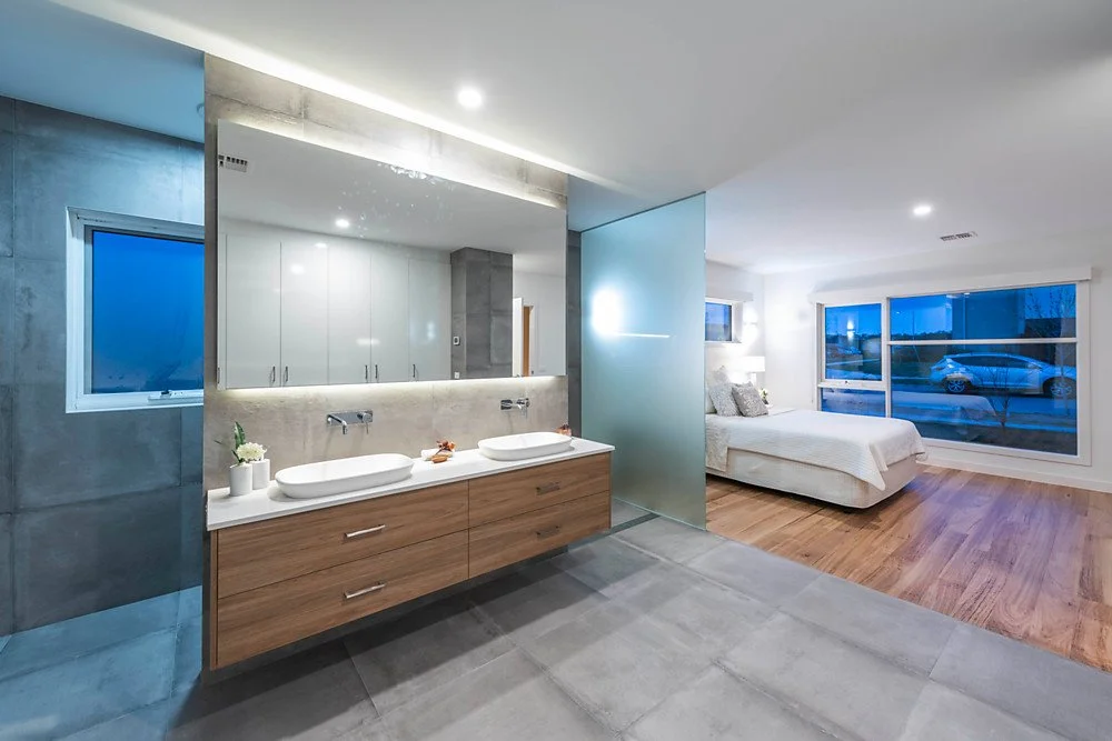 Modern bathroom and bedroom with large window, white bedding, wooden floors, double sinks, and minimalist decor.