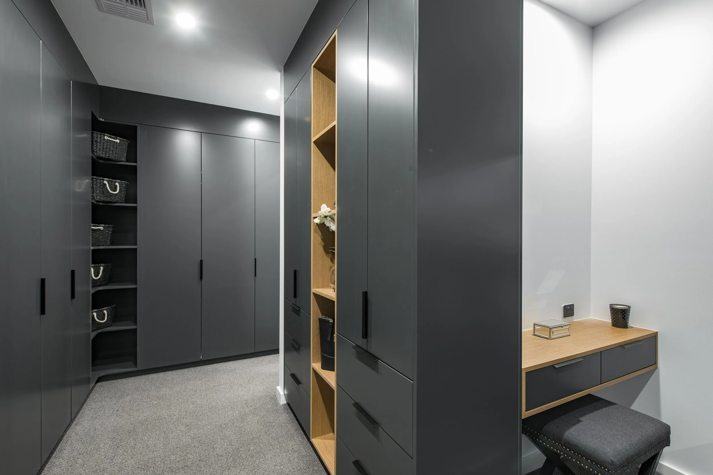 Modern closet with gray cabinets, open shelving, and a small wooden table with a black and gold upholstered bench nearby. The closet features a gray carpeted floor and white walls, with some baskets and decorative items on the shelves.