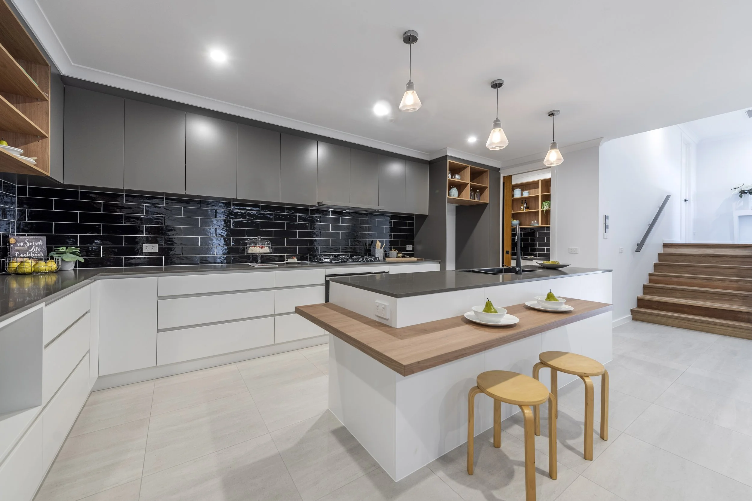 Modern kitchen with white cabinets, black subway tile backsplash, gray upper cabinets, wooden open shelving, and a white island with a wooden countertop. Small stools are at the island, and there are three pendant lights hanging from the ceiling. The