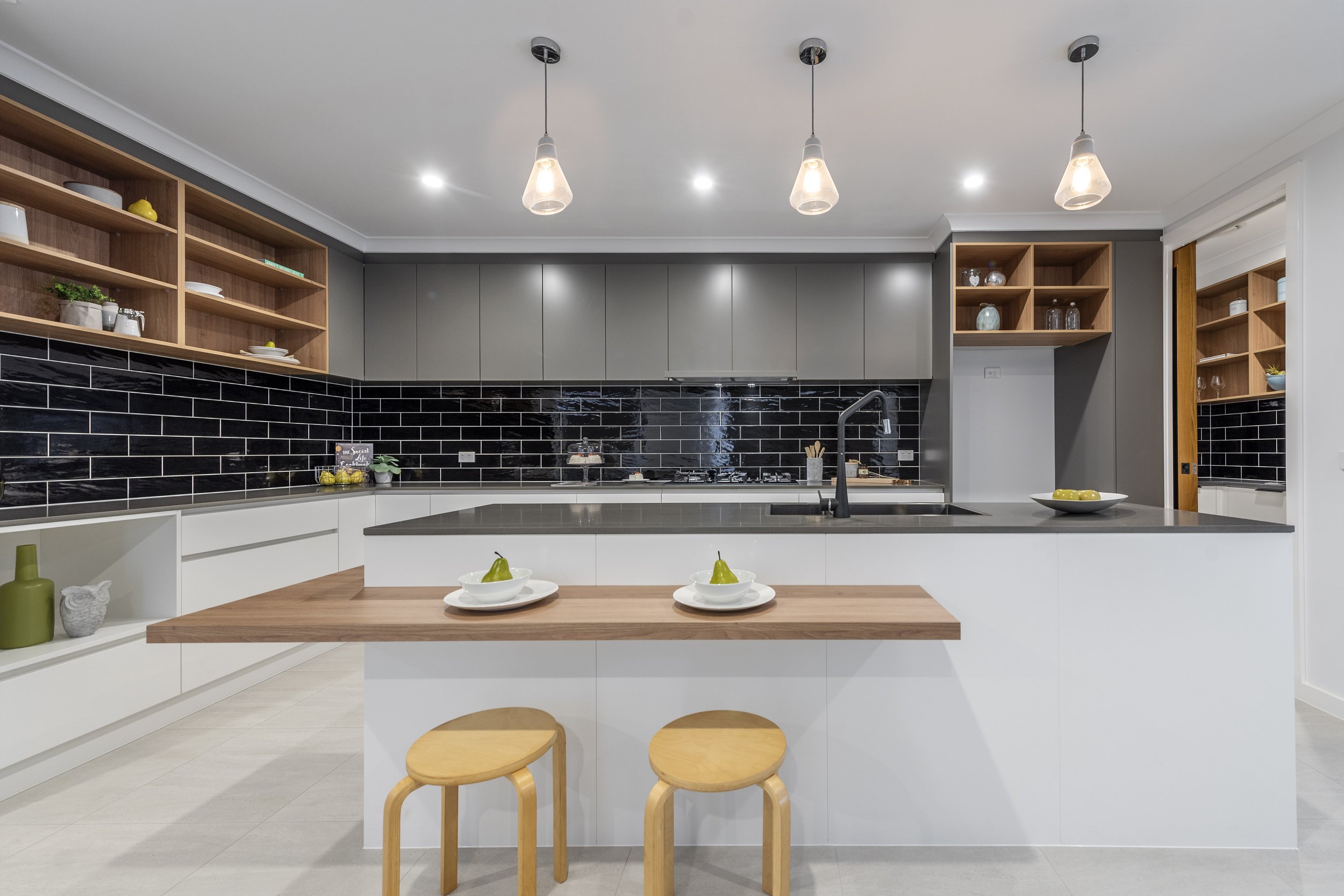 Modern kitchen with open wooden shelves, black subway tile backsplash, gray upper cabinets, white lower cabinets, wooden countertop island, and three pendant lights with glass shades. There are some decorative items on the shelves and a bowl of lemon
