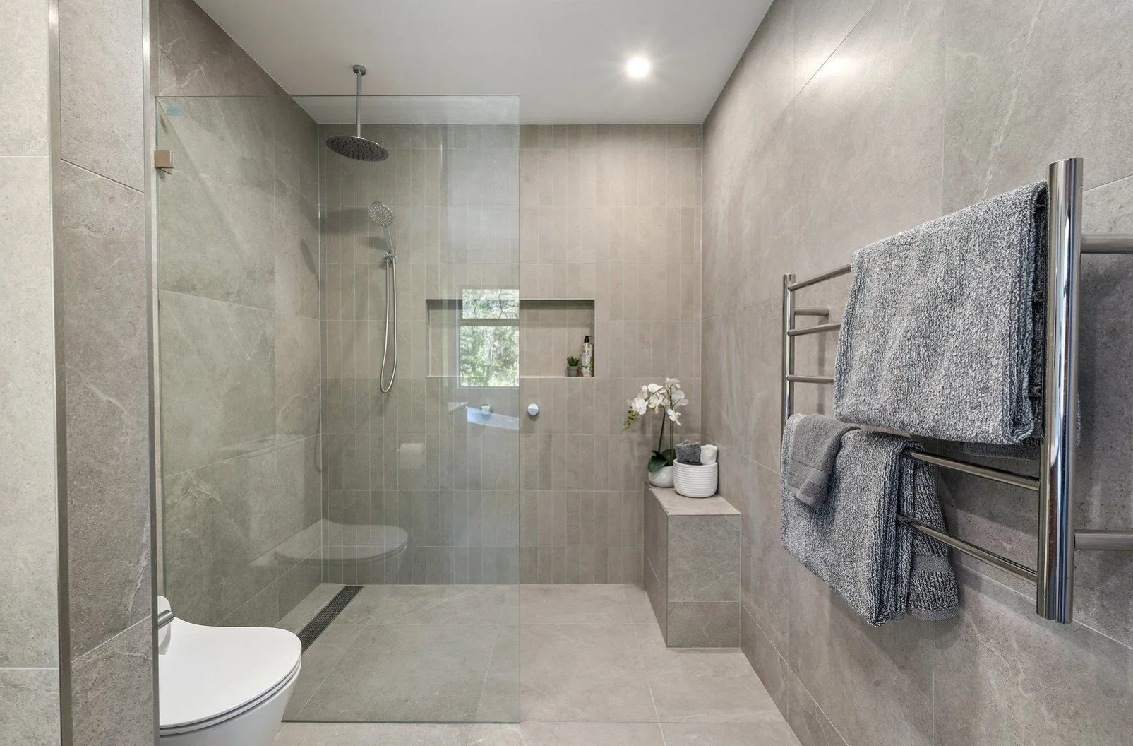 Modern bathroom with walk-in shower, glass enclosure, gray tiled walls and floor, wall-mounted towels rack with gray towels, small ledge with decorative plant and toiletries, and a window with natural light.