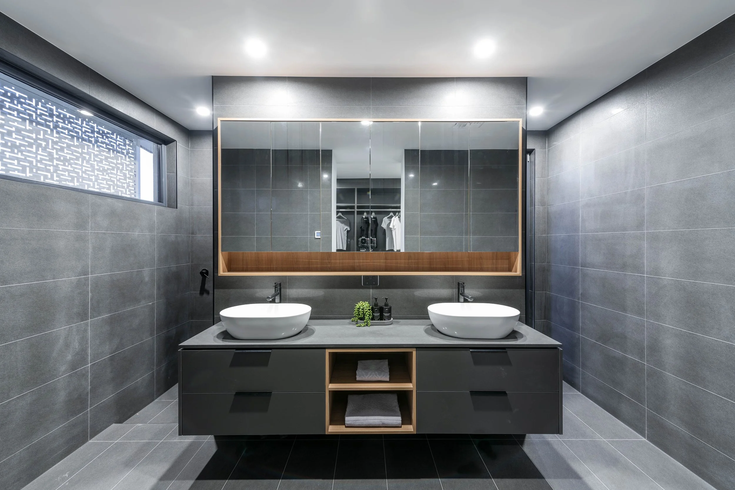 Modern bathroom with dark gray tiled walls and floor, double sinks, large mirror, small window, and contemporary decor.