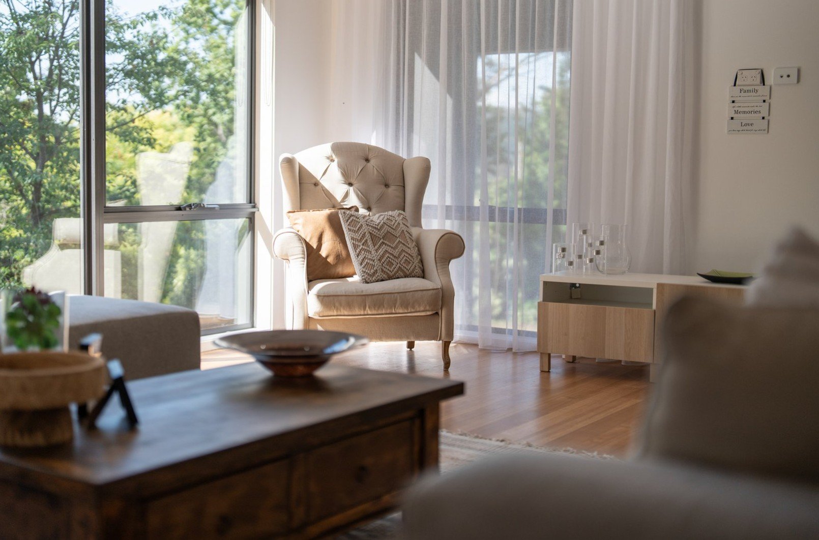 A cozy living room with a beige high-back armchair near large windows with white curtains, decorated with pillows, overlooking green trees outside. There is a wooden coffee table with bowls in the foreground and a white sideboard with glassware on th