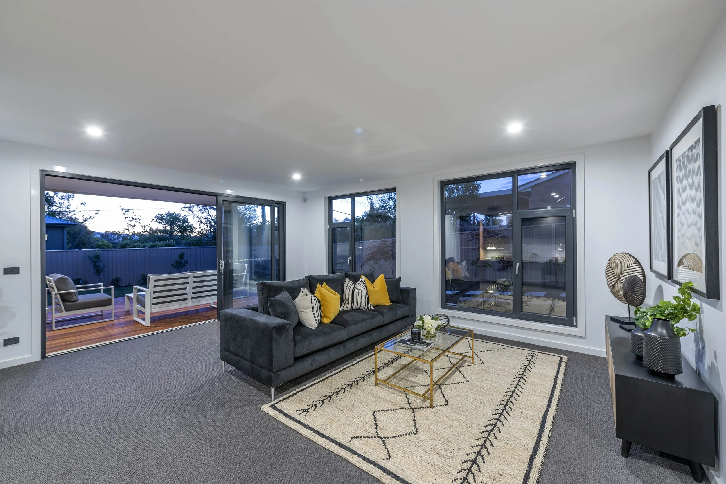 Living room with a black sofa, yellow and white pillows, a glass coffee table with a flower arrangement, a patterned rug, and large windows and sliding doors leading to a wooden deck with outdoor seating, during twilight.