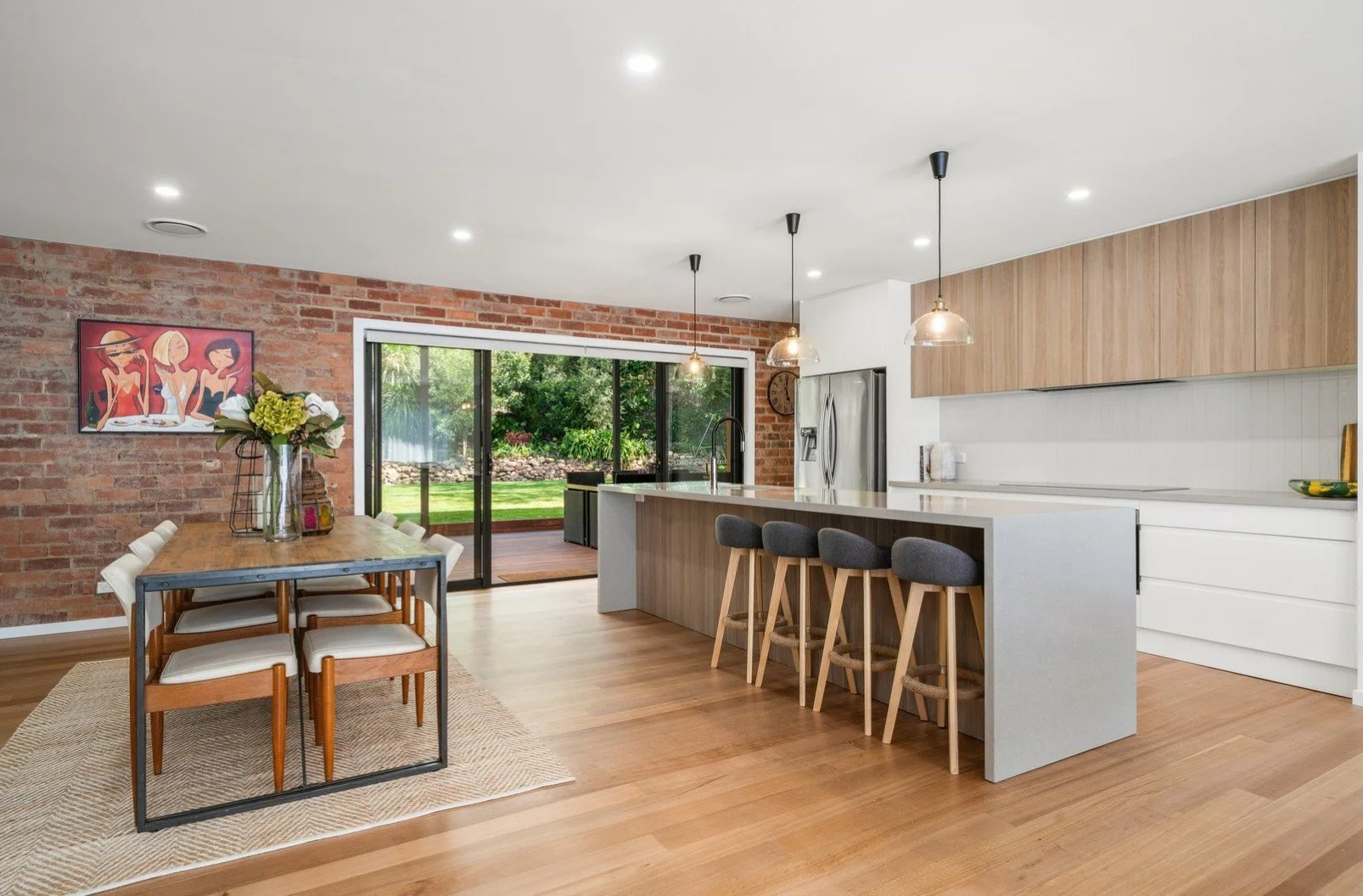 Modern open-concept kitchen and dining area with brick accent wall, large sliding glass door leading to backyard, wooden flooring, and contemporary decor.