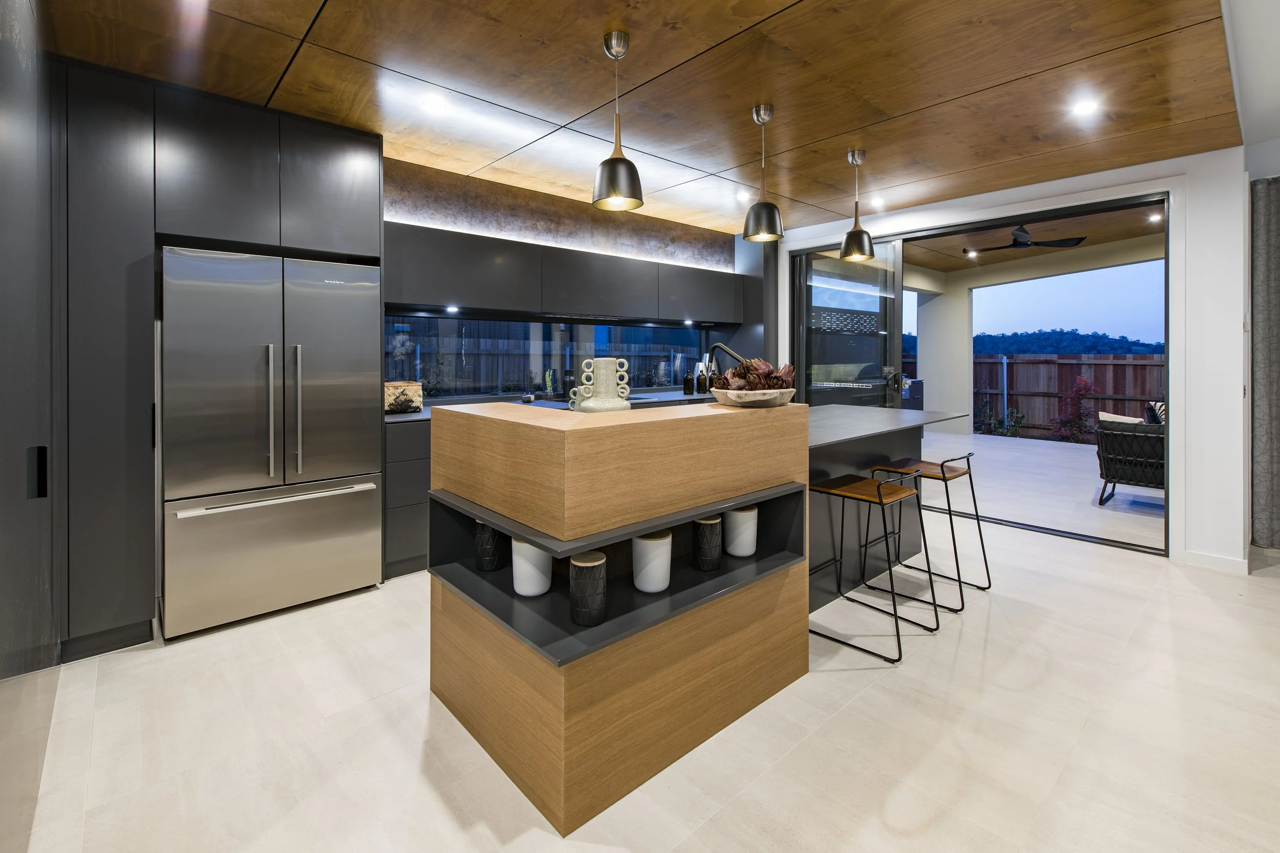 Modern kitchen with black cabinets, a wooden island, stainless steel appliances, pendant lights, and sliding glass doors leading to an outdoor patio with a view of trees and a blue sky.