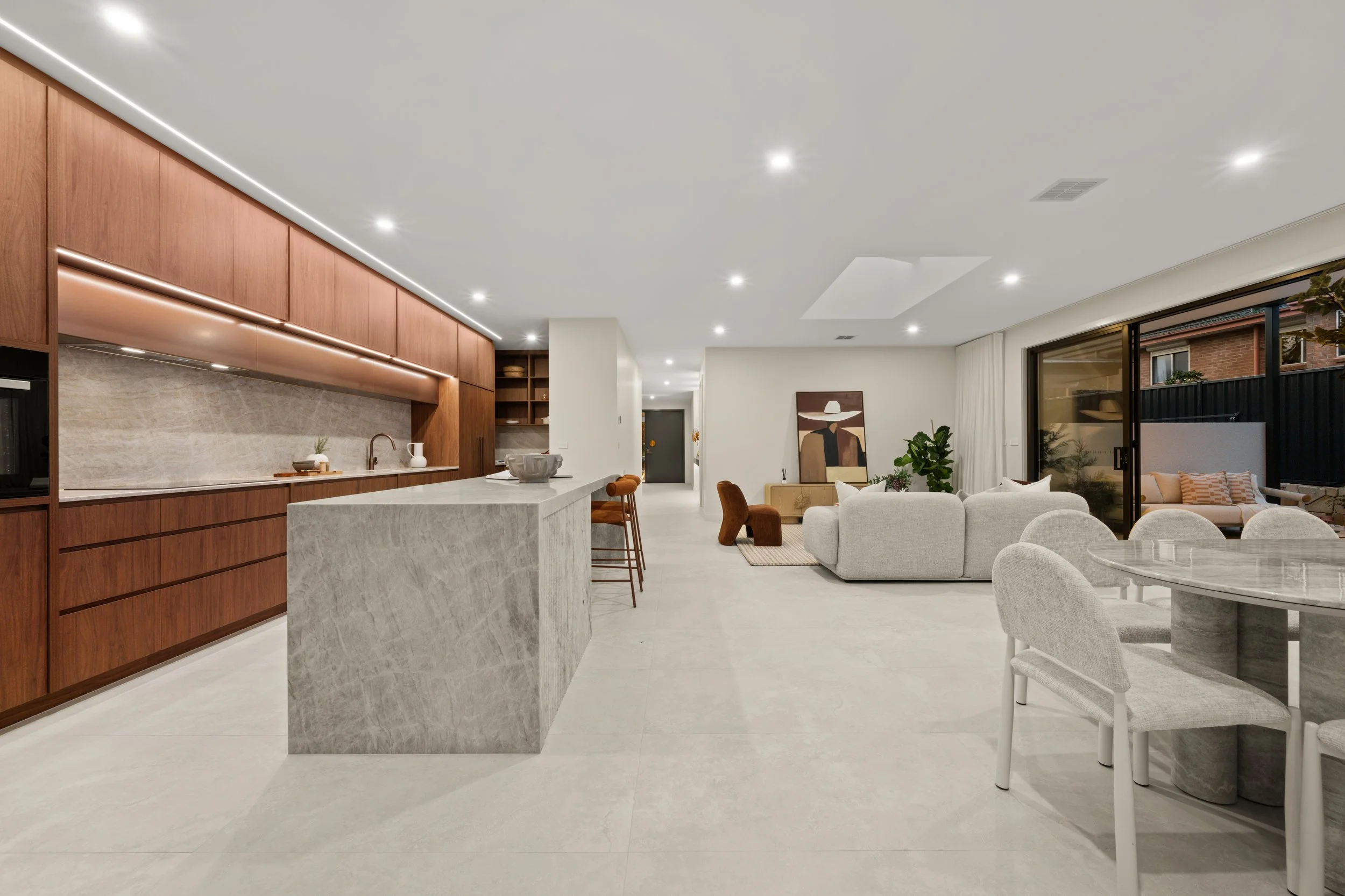 Open-concept living space with a modern kitchen featuring wooden cabinets and marble countertops, a dining area with a marble table and upholstered chairs, and a cozy seating area with a white sofa, artwork, and large sliding glass doors leading outd