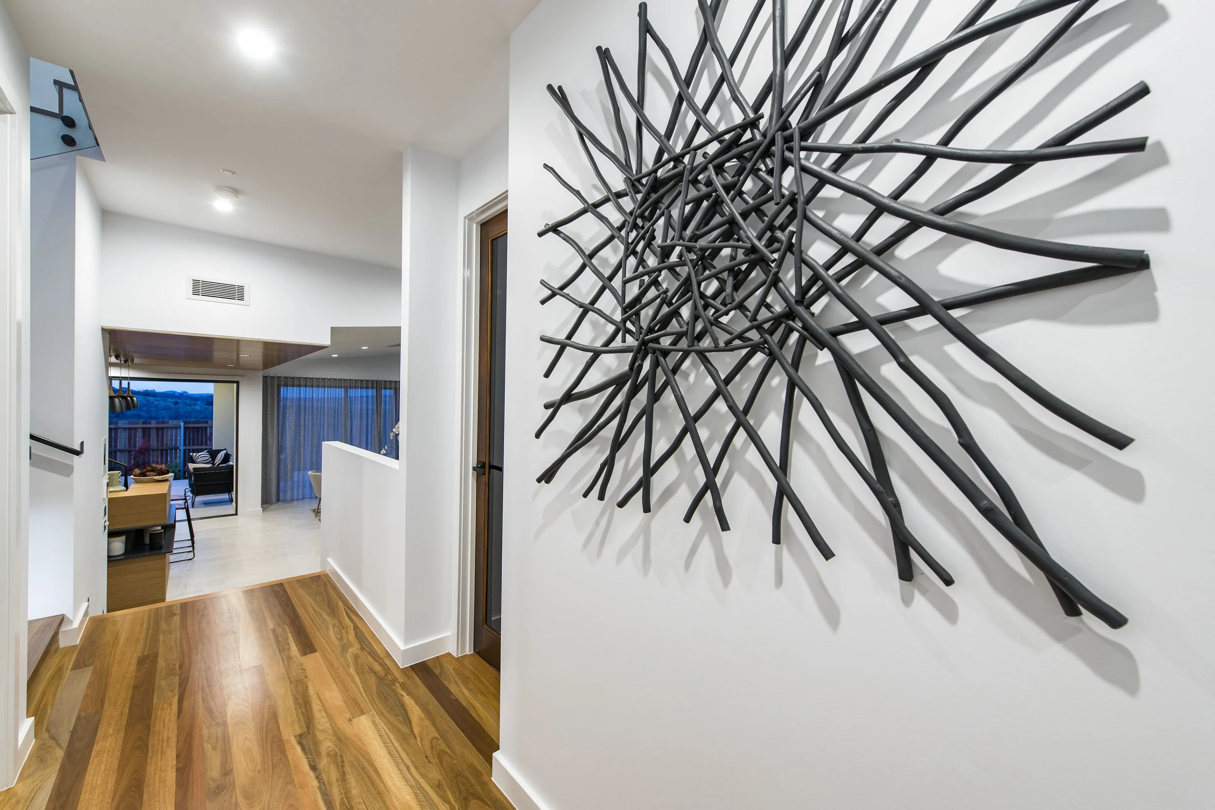 Modern interior hallway with wooden flooring, white walls, and unique black metal wall art resembling intertwined sticks, with a view into the living room area which has large windows with curtains and outdoor scenery.