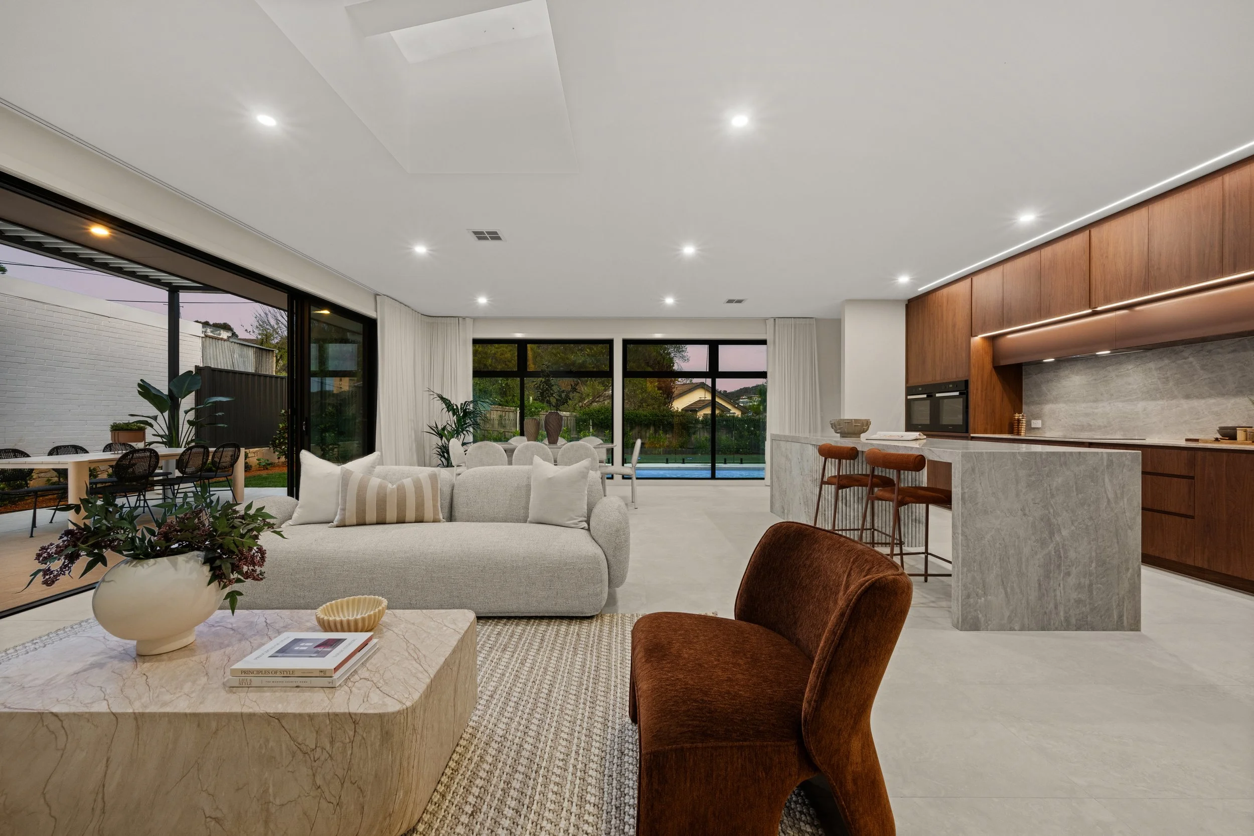 Modern living room with a white sofa, brown armchair, marble coffee table with books, potted plants, large windows, and a kitchen area with wooden cabinets and bar stools.