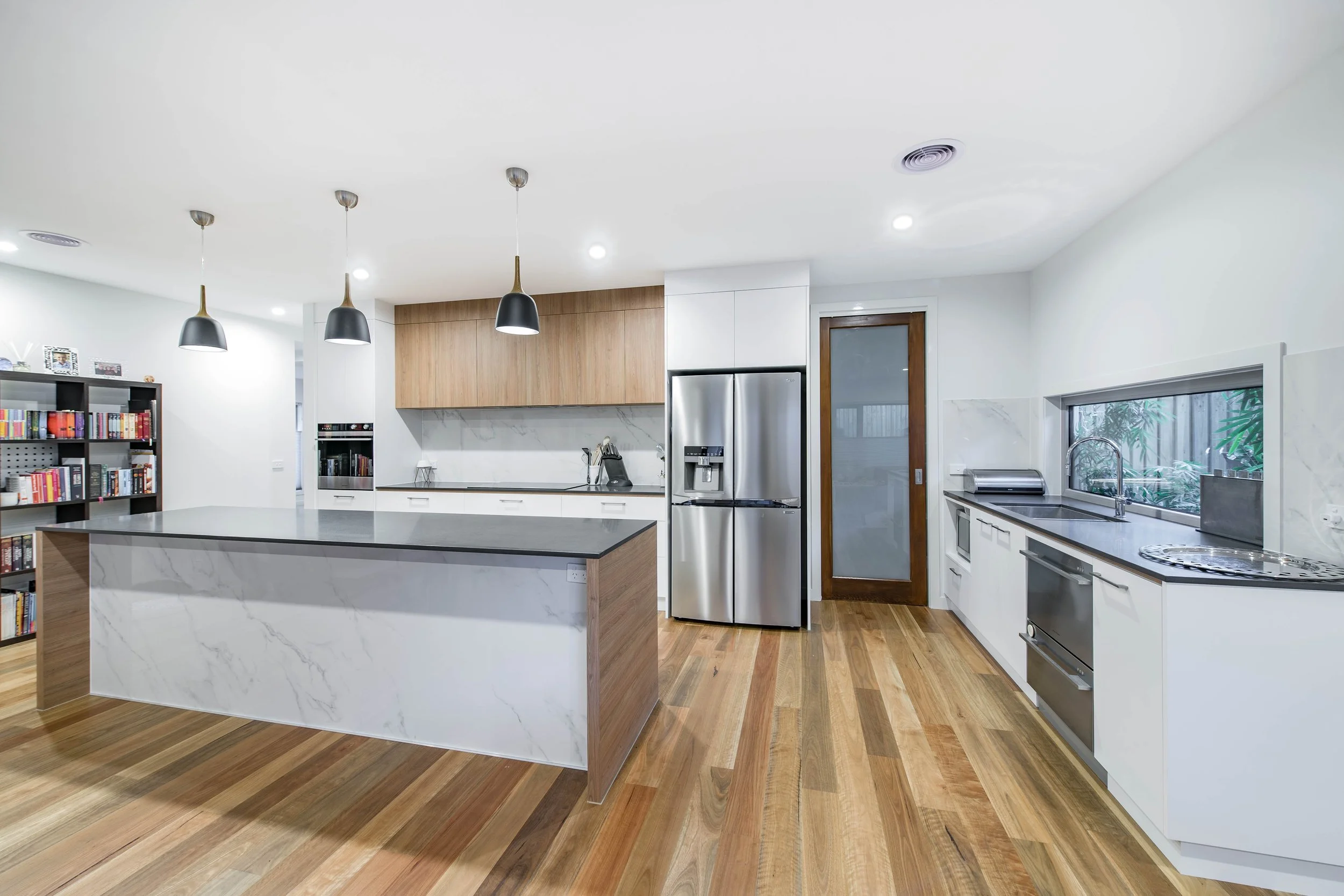 Modern kitchen with white cabinets, stainless steel refrigerator and oven, marble island, wooden flooring, large window with plants outside, black pendant lights, bookshelf on the left.