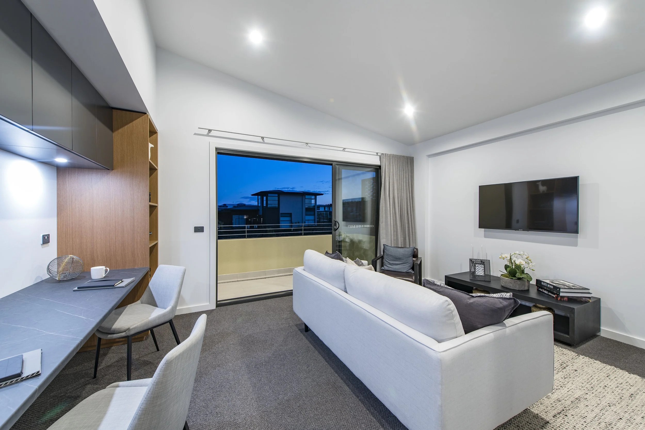 Modern living room with white sofa, black side table with flower arrangement, wall-mounted television, glass sliding door showing buildings outside, beige curtains, and dining area with chairs and notebooks on a gray table.