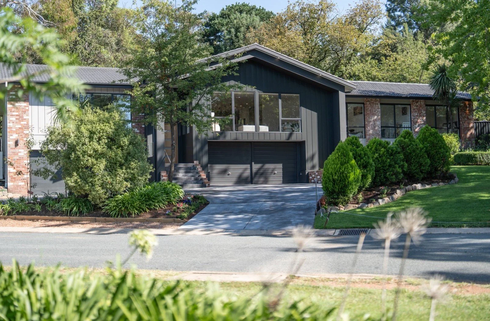 Modern house with black siding and brick accents, surrounded by green trees and shrubs. The house has a two-car garage, stairs leading to a second-floor balcony, and a well-maintained lawn.