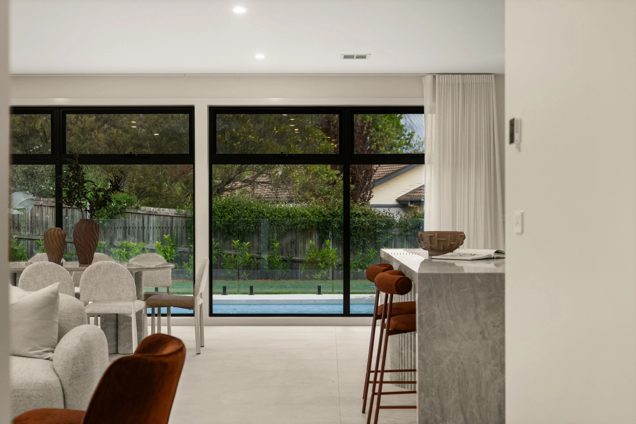 Interior view of a modern living space with large windows overlooking a backyard with trees and shrubs, dining table with chairs, and a kitchen counter with decorative bowls and books.