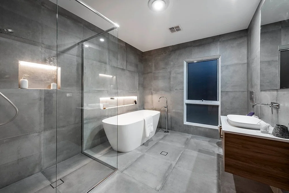 Modern bathroom with gray tiled walls and floor, a freestanding bathtub, a walk-in shower with glass enclosure, a window, and a wooden vanity with a vessel sink.