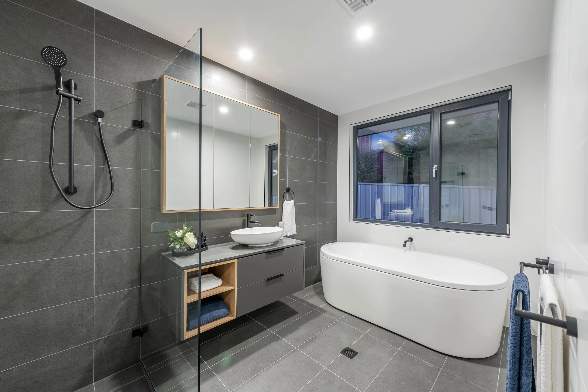 Modern bathroom with dark gray tiled wall, a white bathtub, a sink with a gray cabinet, a large window, and a glass shower partition.