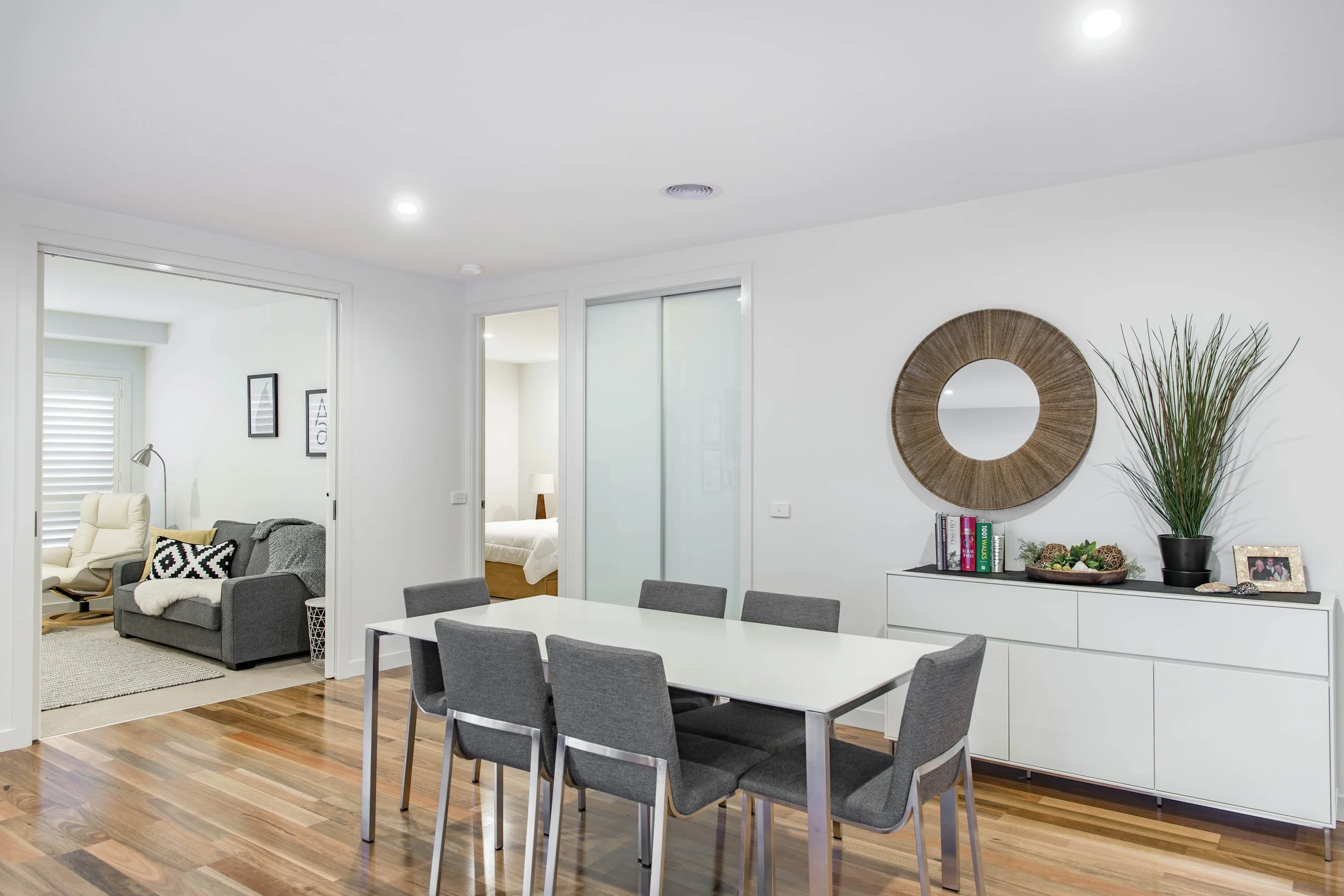 Open-plan living and dining room with white walls, wooden flooring, a white dining table with six gray chairs, a white sideboard decorated with books, plants, and framed photos, a round mirror on the wall, a corner with decorative grass, and a visibl
