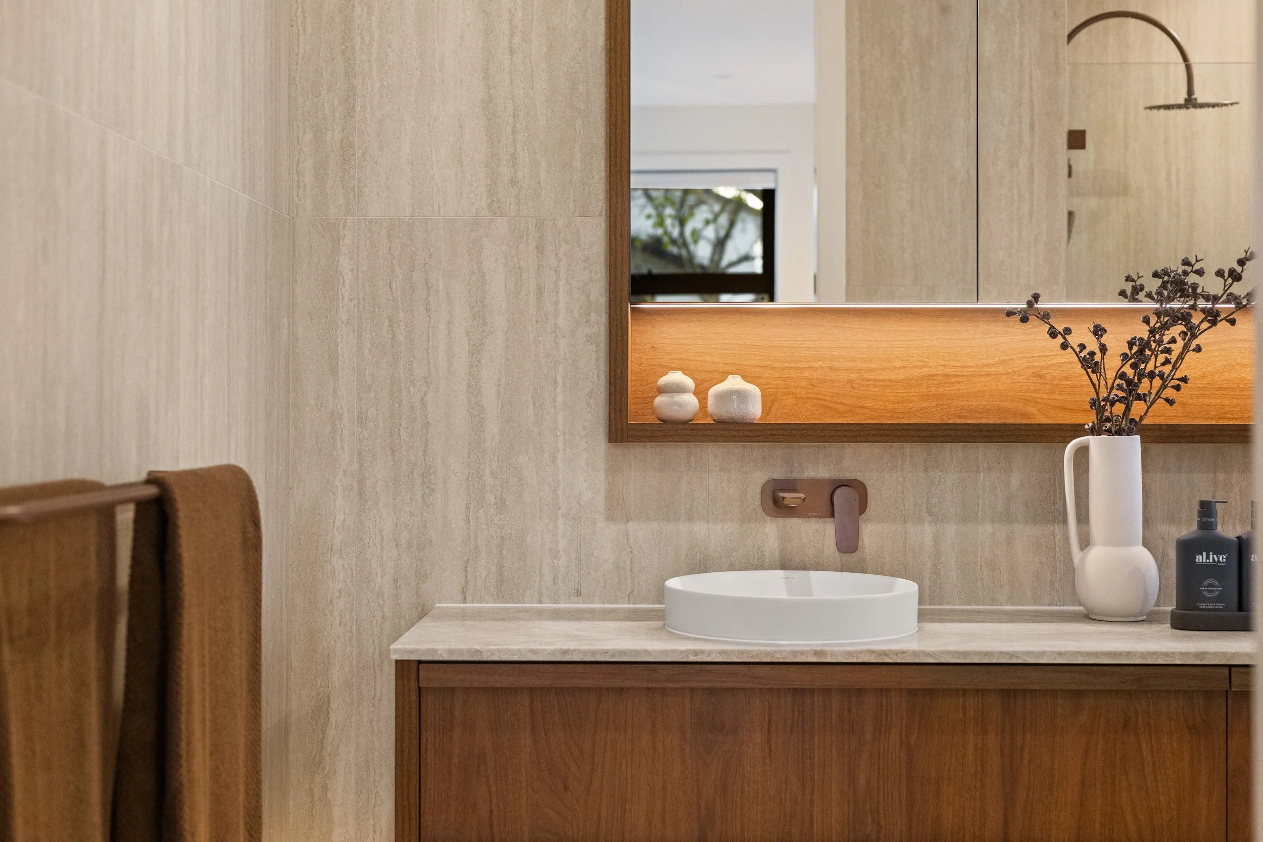 A bathroom vanity with a round white sink, a mirror, a wooden framed mirror, a beige countertop, a white vase with dried purple flowers, brown towels on a wooden towel rack, and various soap bottles.