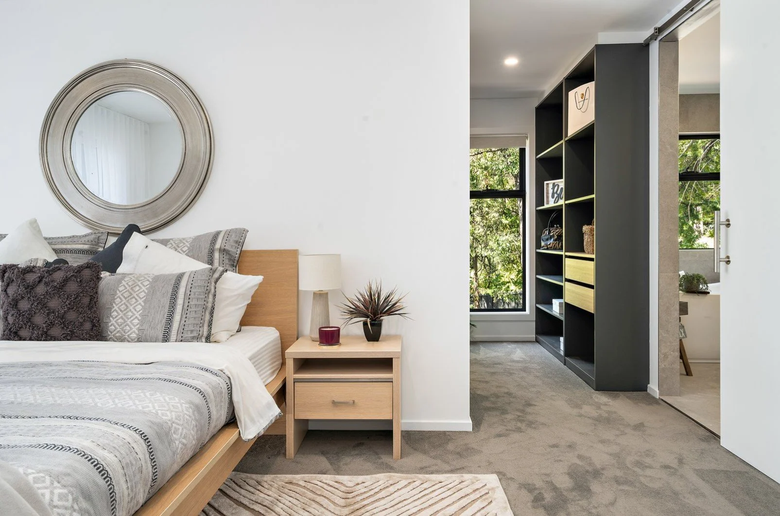 A bedroom with a bed, nightstand, lamp, and decor. The room has a large circular mirror above the bed, and natural light coming through the window.