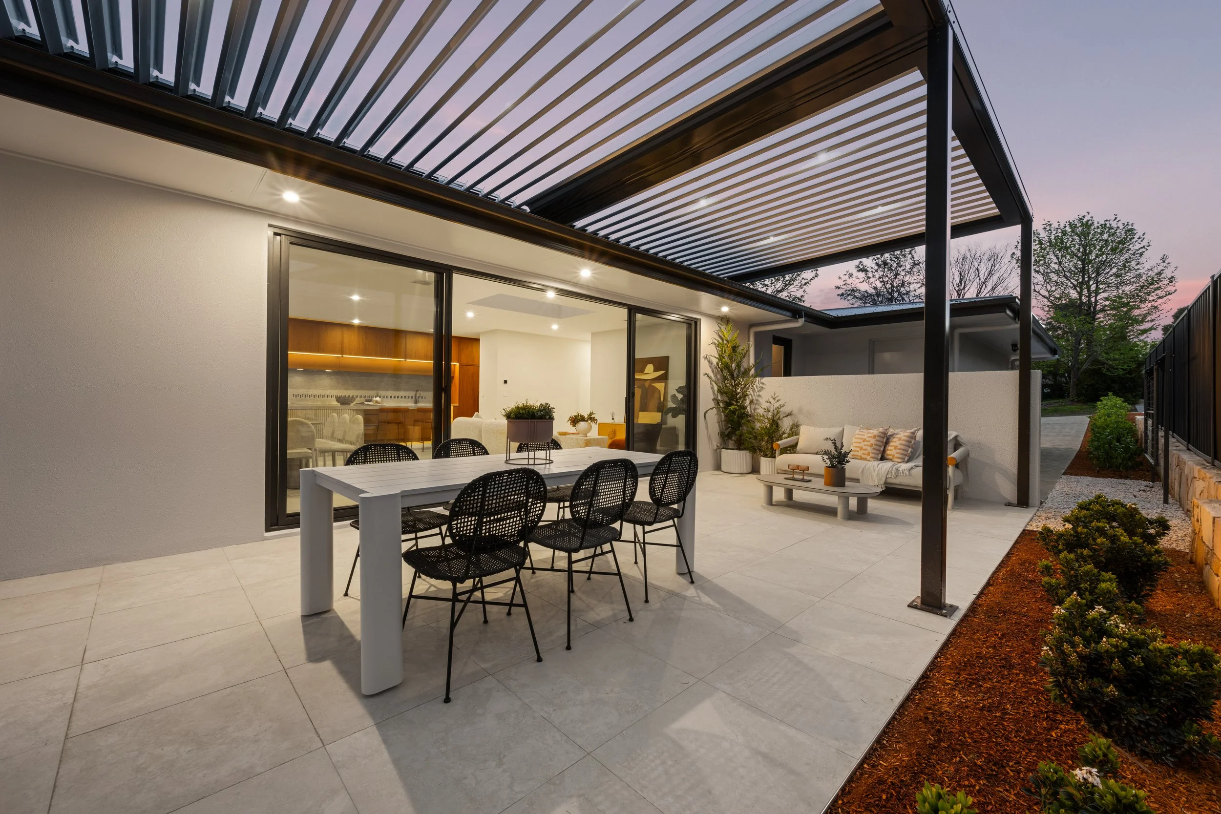 Outdoor patio with dining table and chairs, sofa, plants, and a slatted pergola at sunset.