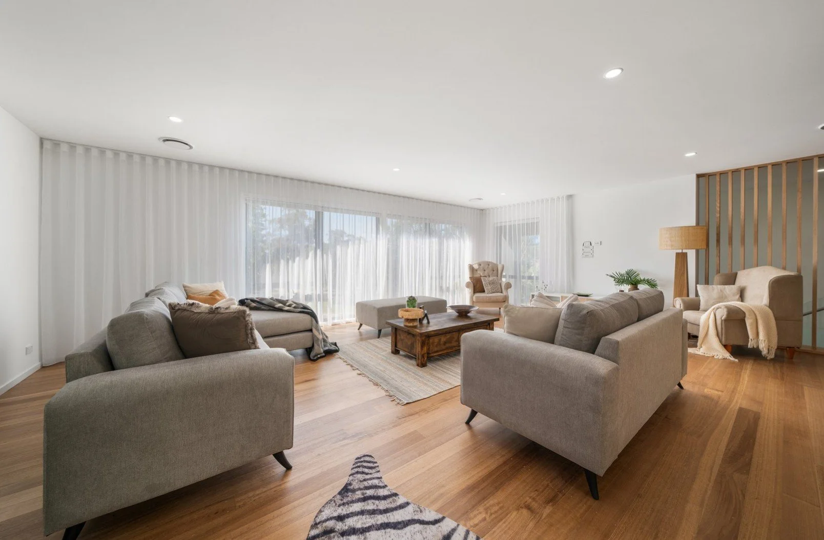 Spacious living room with beige sofas and armchair, wooden coffee table, area rug, large windows with sheer curtains, hardwood floor, potted plants, floor lamp, and minimalist decor.