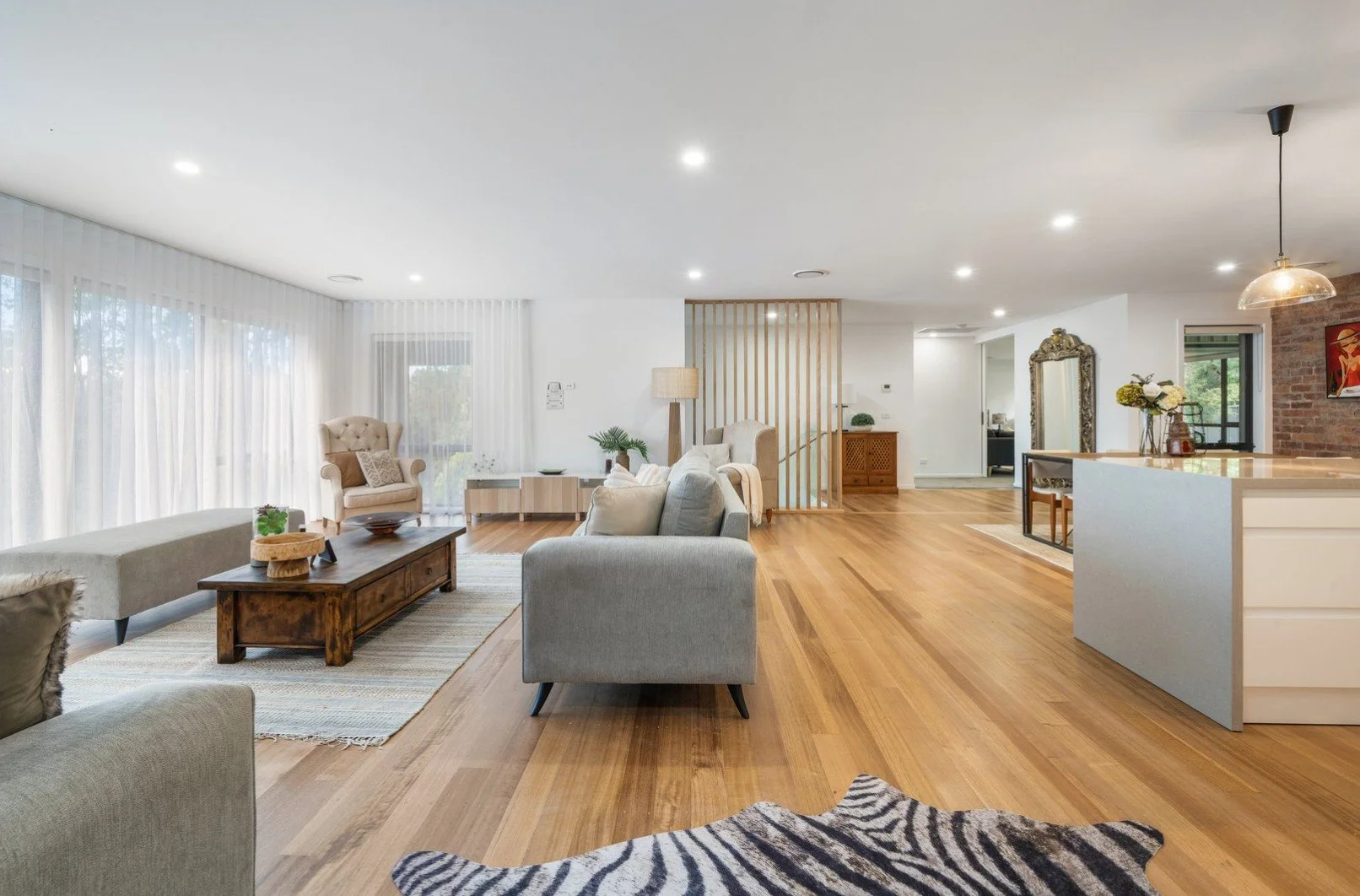 Spacious, brightly lit living room with wooden floors, light-colored furniture, large windows with sheer curtains, and decorative accents.