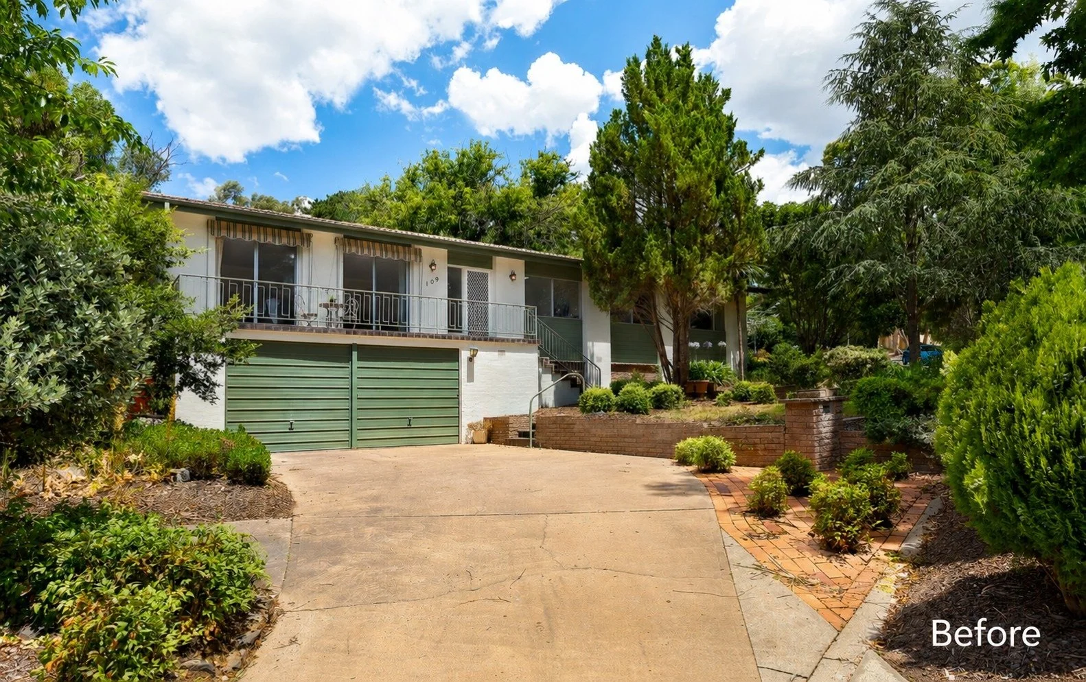 The front view of a house with a driveway, surrounded by greenery and trees, with a bright blue sky with white clouds above.