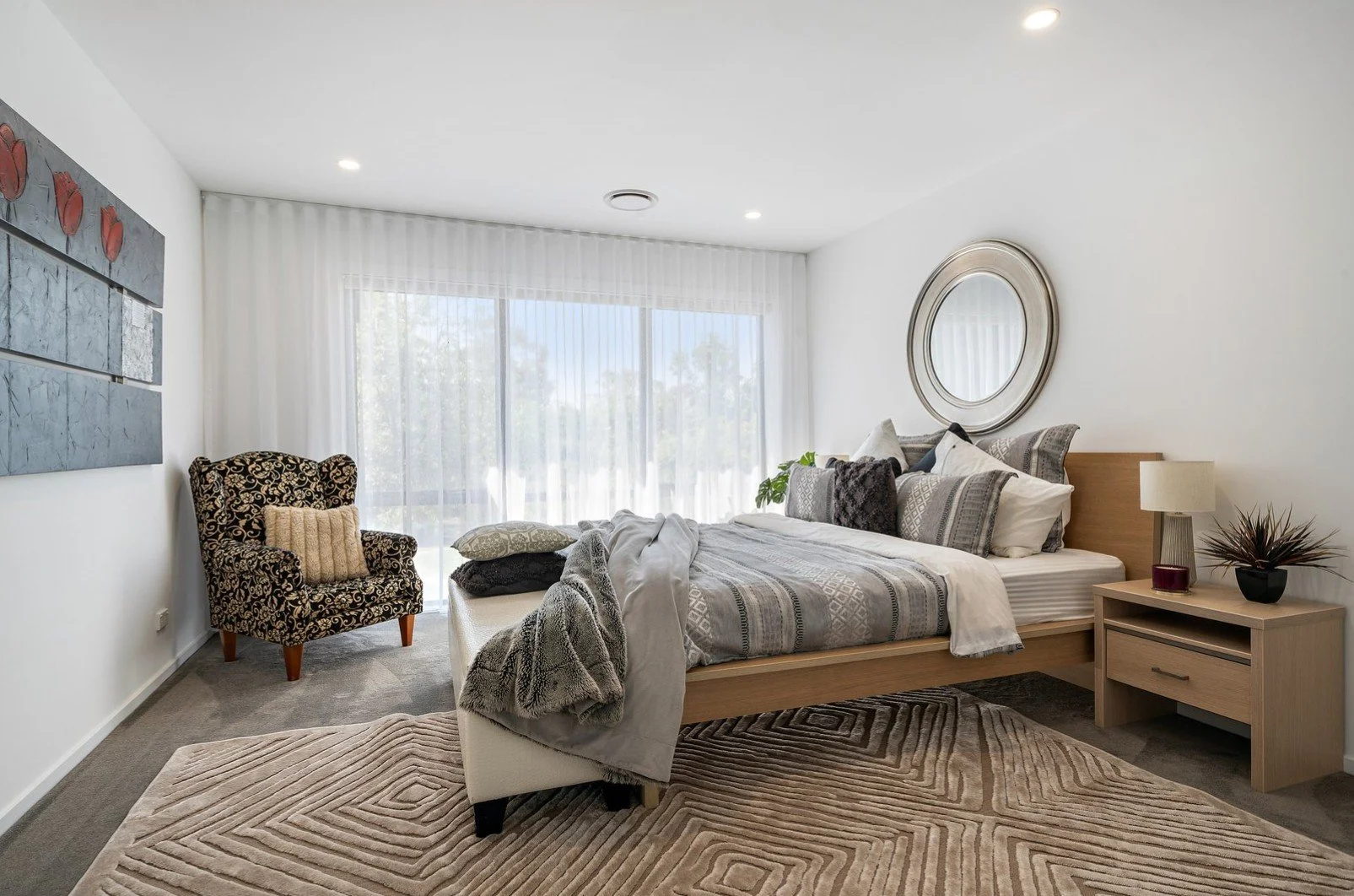 A bedroom with a wooden bed, nightstand, and mirror, featuring patterned pillows, a lamp, and artwork, with a large window and sheer curtains.