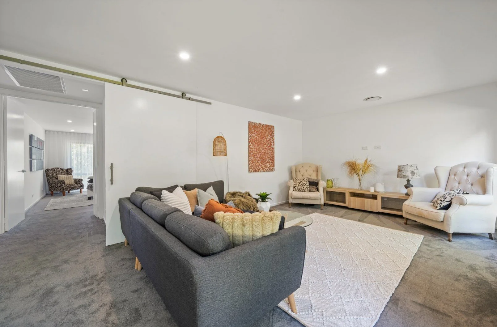 Living room with gray and white sofas, white armchair, coffee table, area rug, and decorative pillows. Additional seating visible in adjacent room.