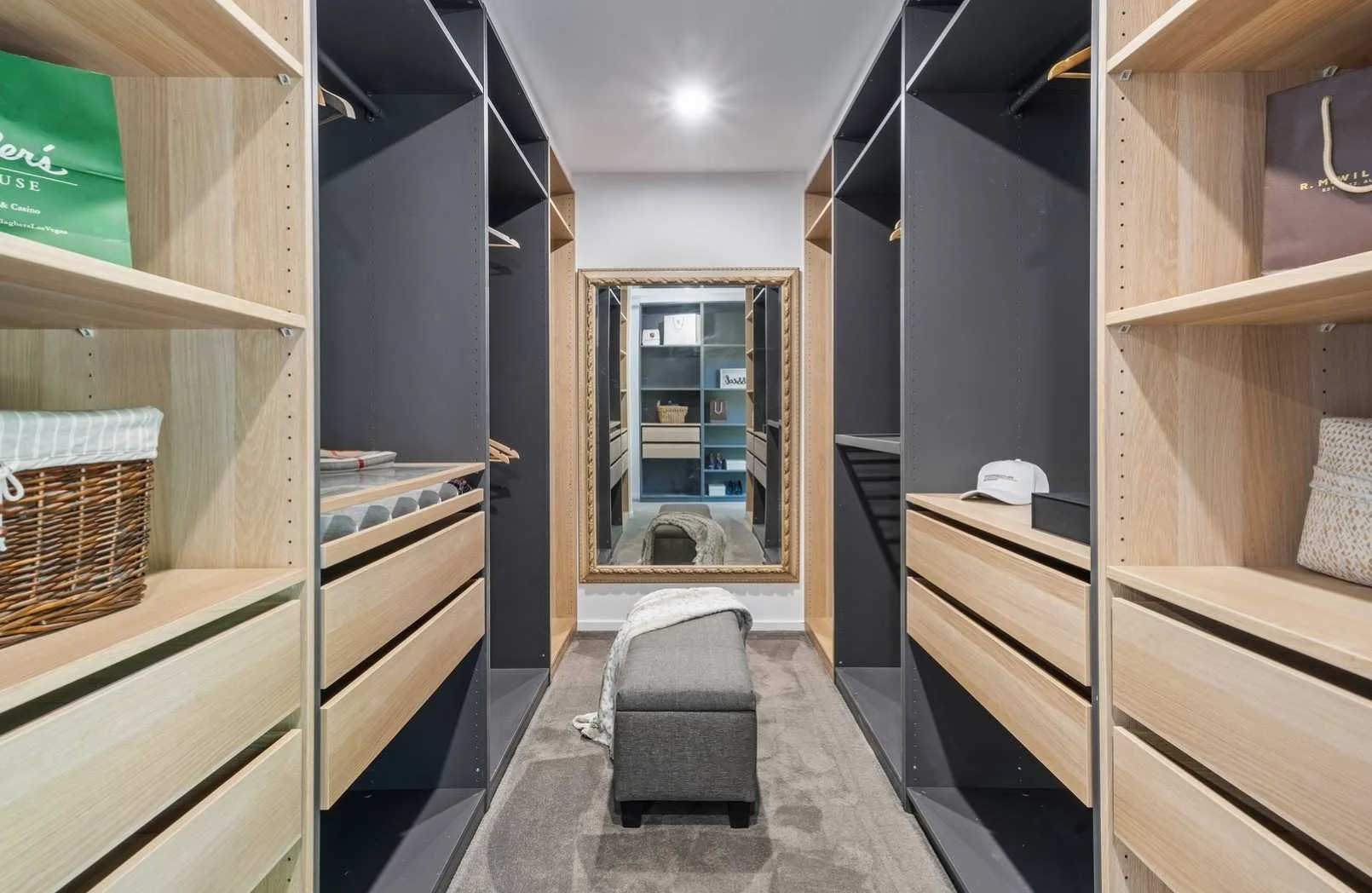 A walk-in closet with wooden and black shelving on both sides, a large ornate mirror at the end, a cushioned bench in front of the mirror, and various personal items like handbags, a hat, and clothing.