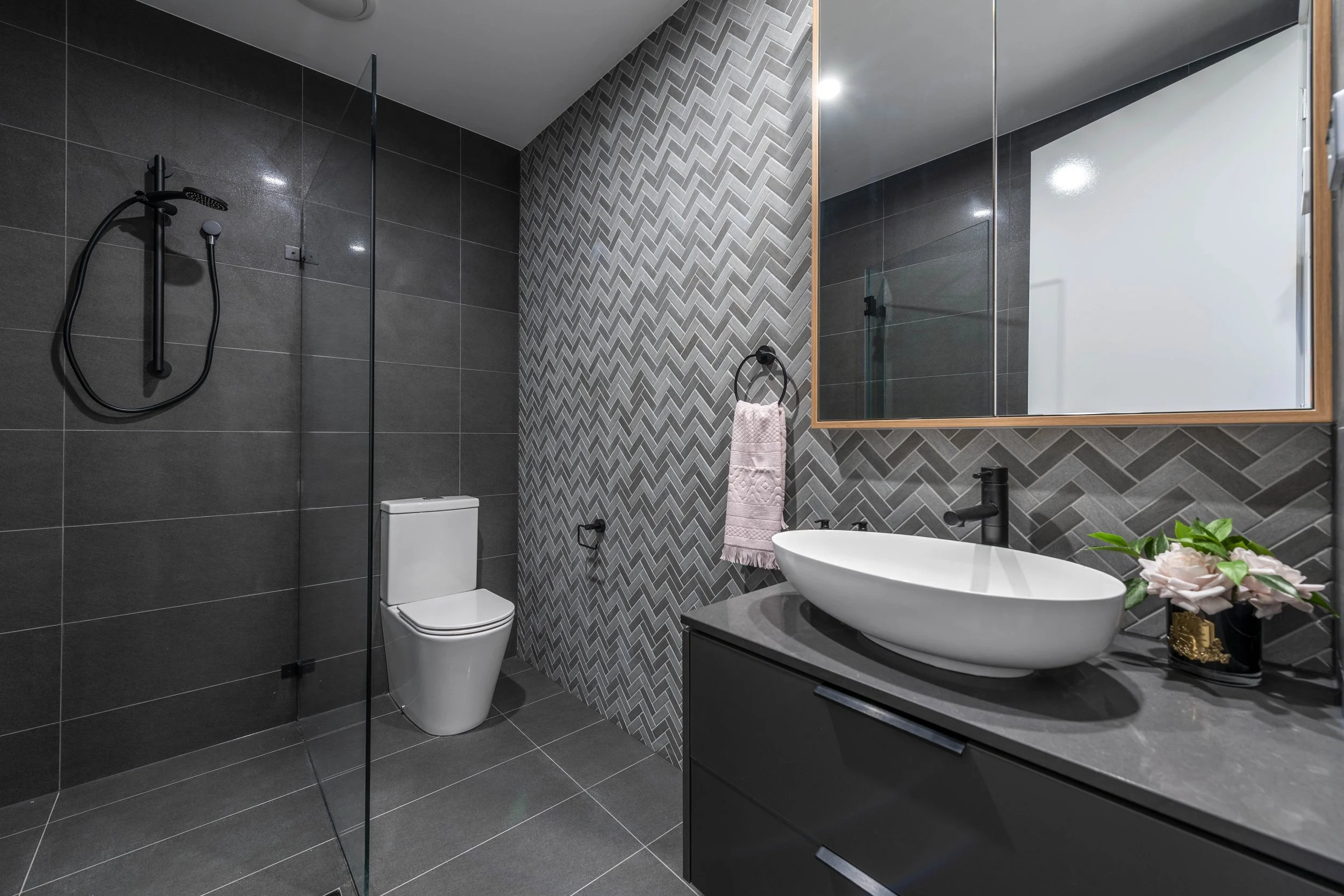 Modern bathroom featuring dark gray tiles, a wall with herringbone-patterned gray tiles, a white toilet, a round vessel sink on a dark vanity, a large mirror, black fixtures, a towel ring with a pink towel, and a plant with pink flowers.