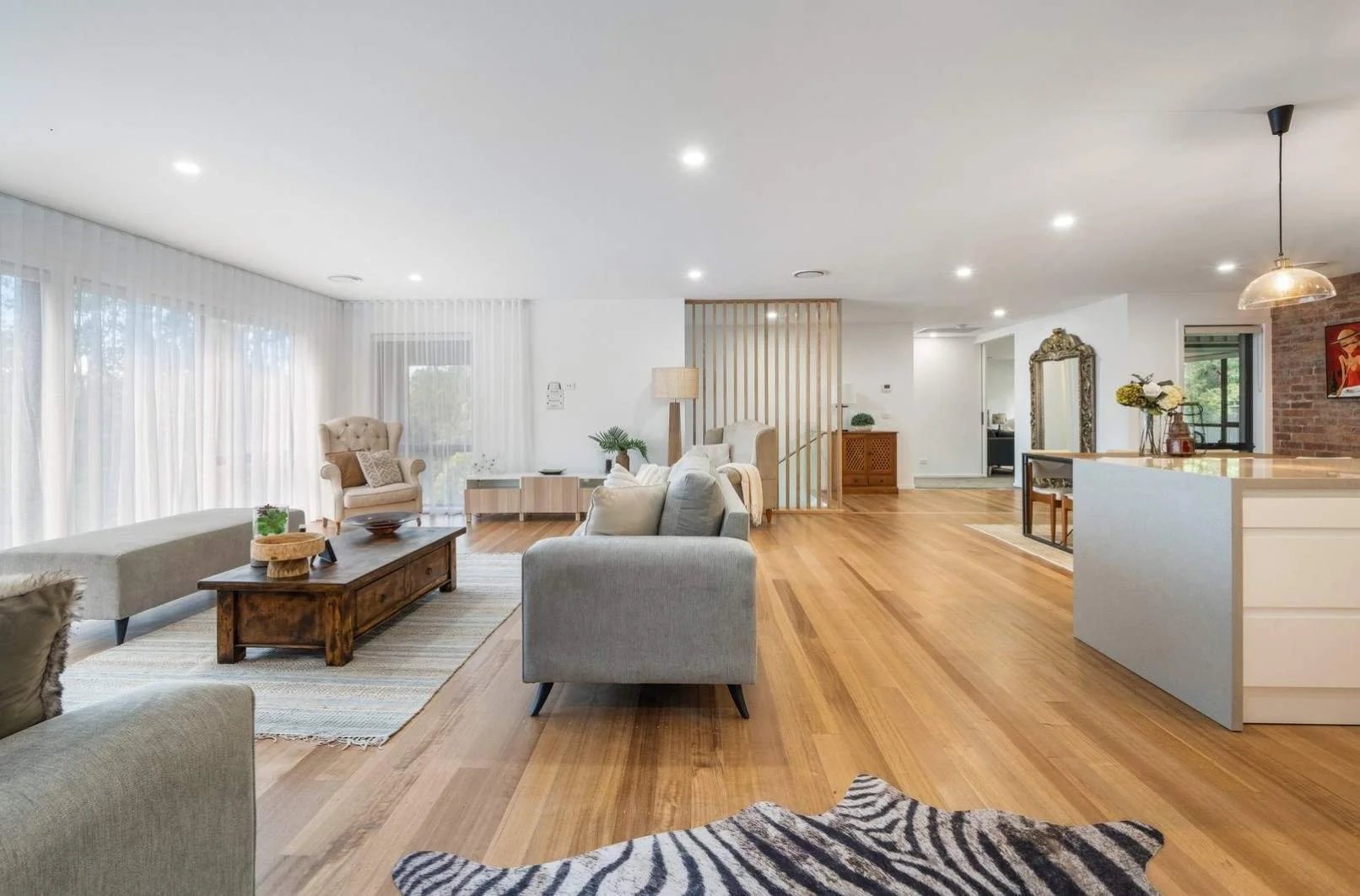 Spacious, brightly lit living room with wooden floors, light-coloured furniture, large windows with sheer curtains, and decorative accents.