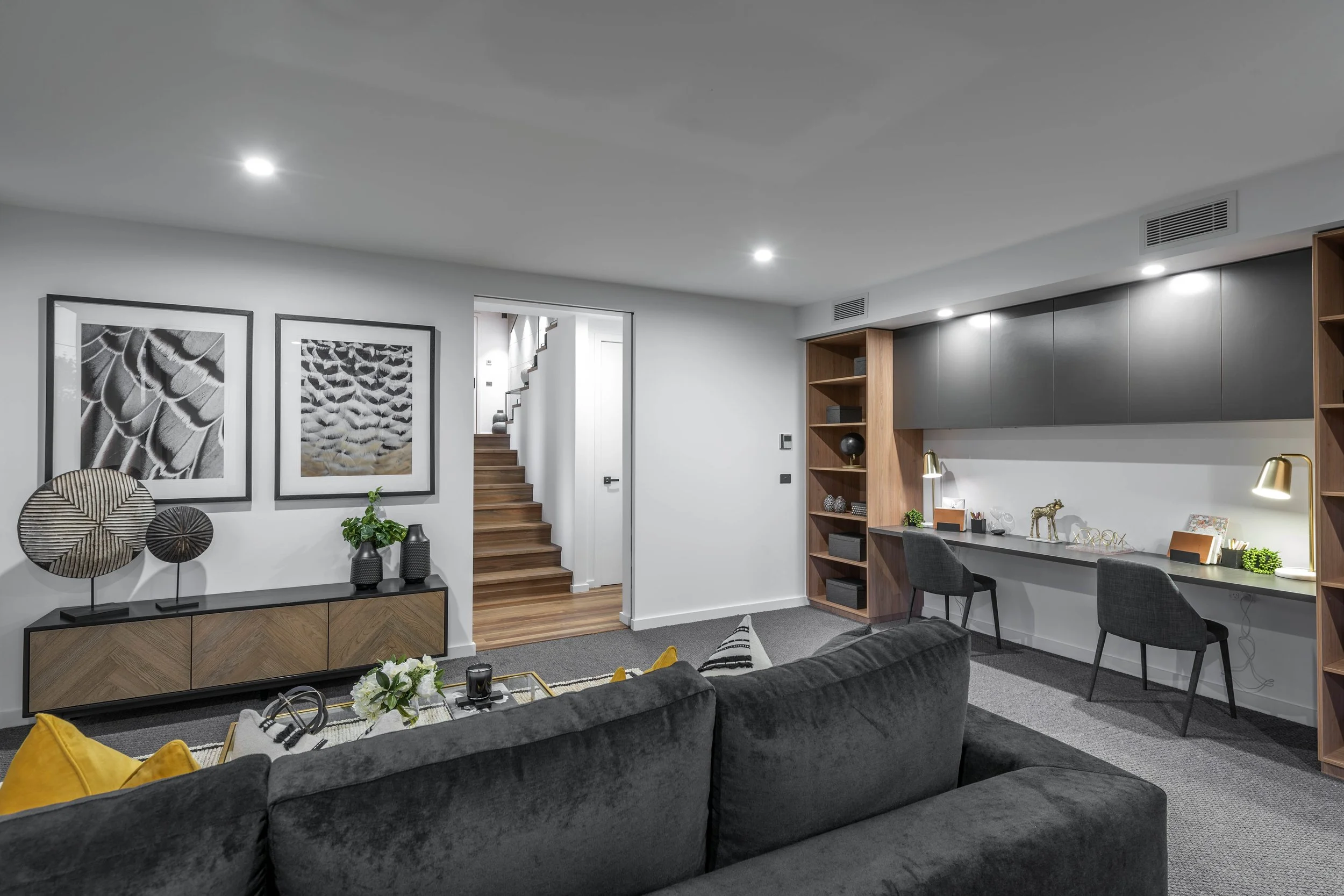 Modern living room with wall art, a black sofa with yellow pillows, a coffee table with flowers, and a workspace with black chairs, wooden shelves, and framed art.