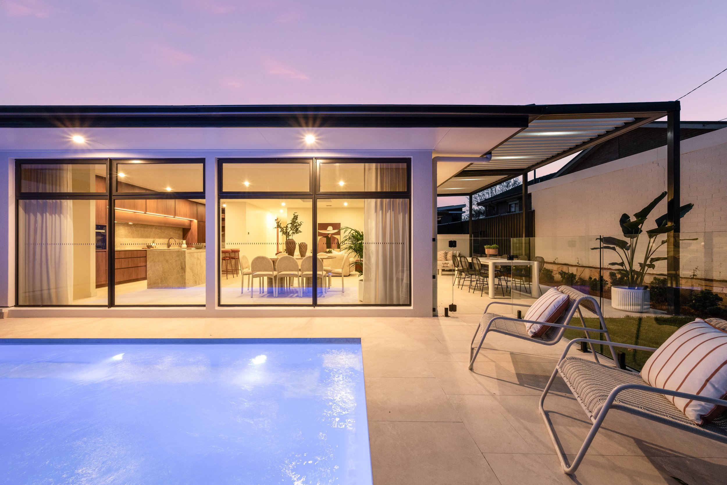 A modern house with a glass wall, open interior featuring a kitchen and dining area, and an outdoor patio with seating, a pool, and landscaping at dusk.