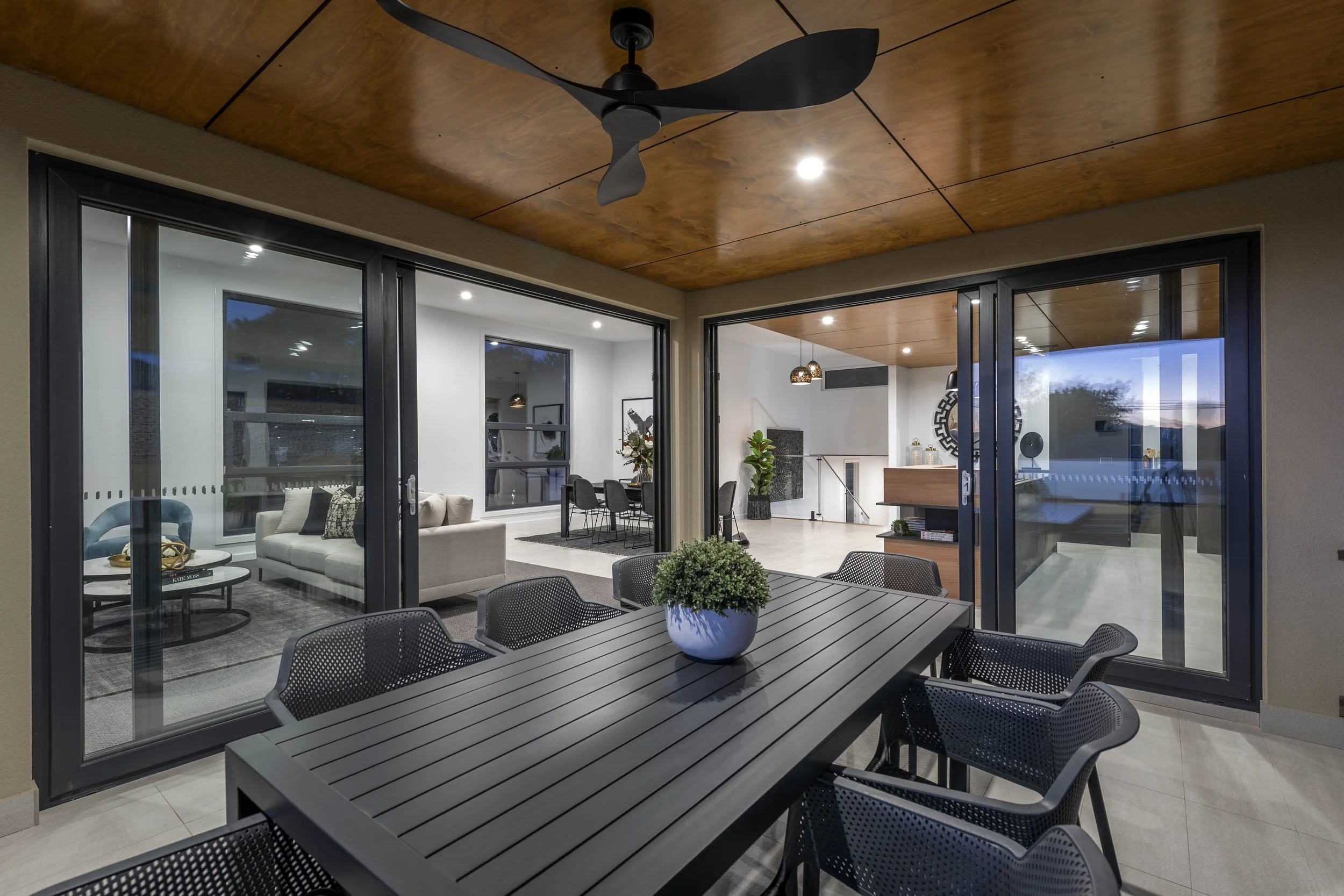 View of a modern indoor-outdoor space with a ceiling fan, sliding glass doors, and a patio dining area with black chairs and a table with a potted plant, leading into a living room and kitchen area.