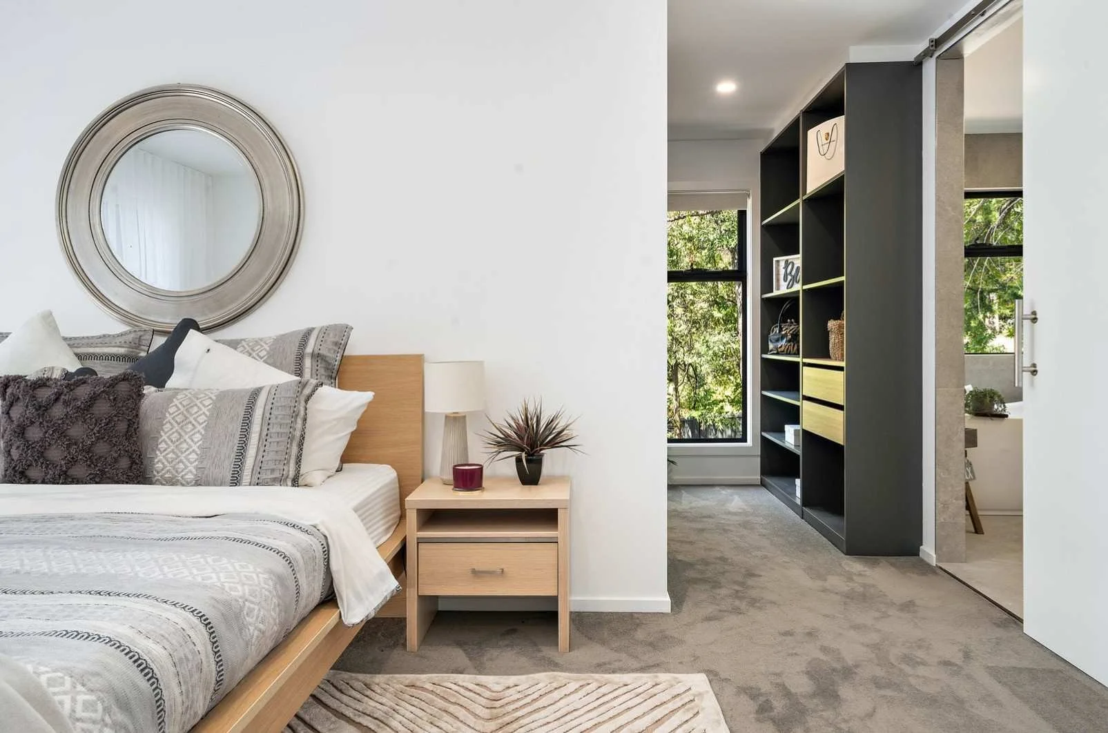 A bedroom with a bed, nightstand, lamp, and decor. The room has a large circular mirror above the bed, and natural light coming through the window.