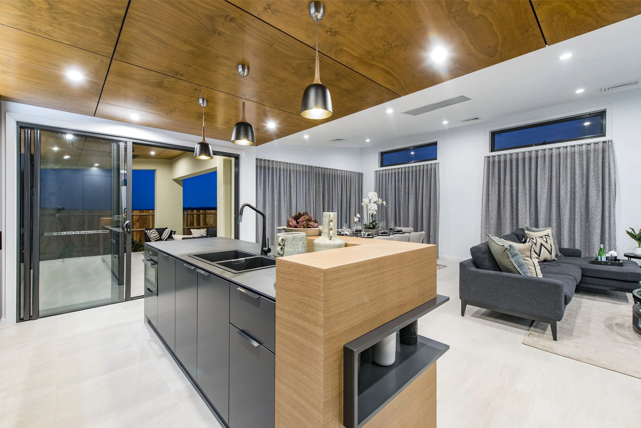Modern open-concept living space with a kitchen island, wooden ceiling with pendant lights, large windows with curtains, and cozy seating area with gray sofa and plants.