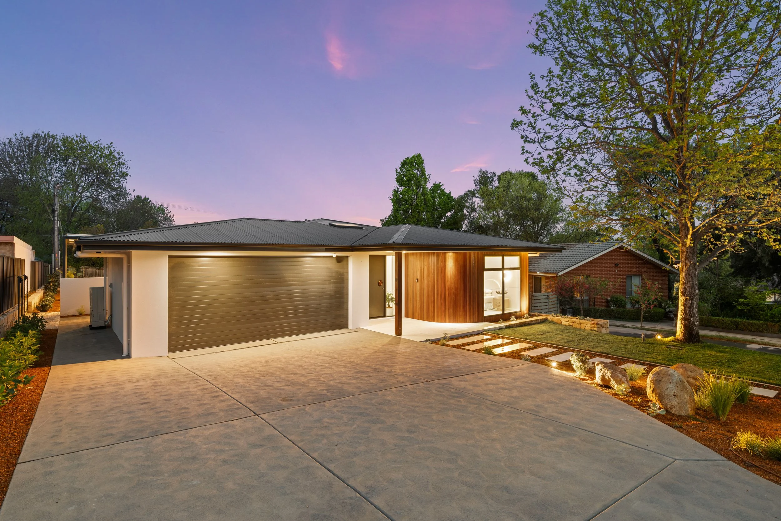 Modern house with a garage and front porch, illuminated by outdoor lighting, surrounded by landscaped yard with rocks and plants, during early evening with a purple sky and trees in the background.