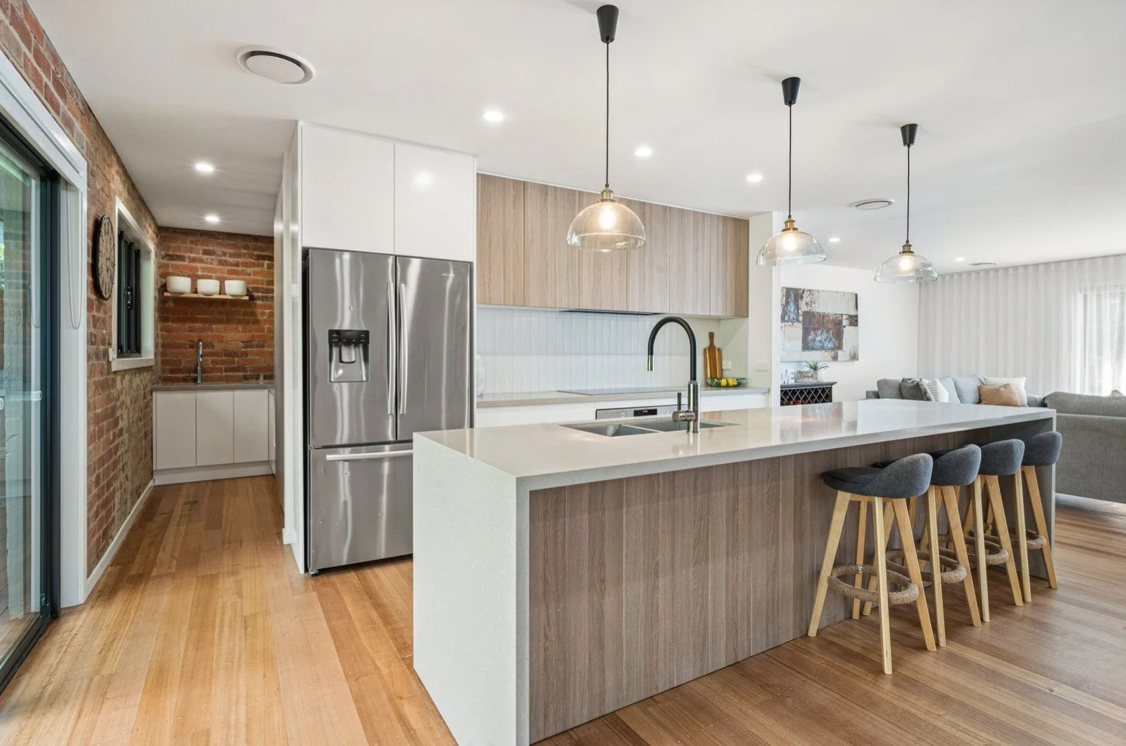 Modern kitchen with white and wood cabinetry, stainless steel refrigerator, island with a sink, gray bar stools, and a living area with a sofa and art on the wall.