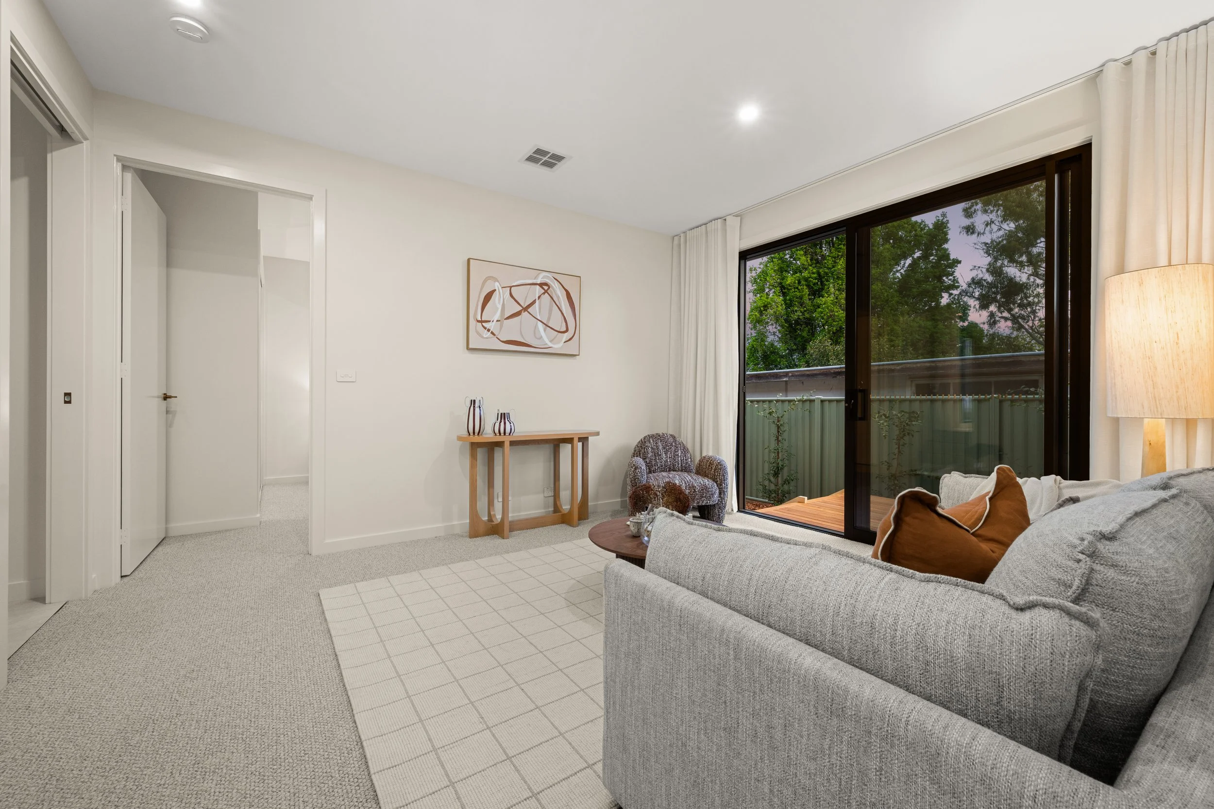 Living room with a large window, beige couch with brown pillows, a wall art, a small wooden table, an armchair, and a floor lamp, with a view of greenery outside.
