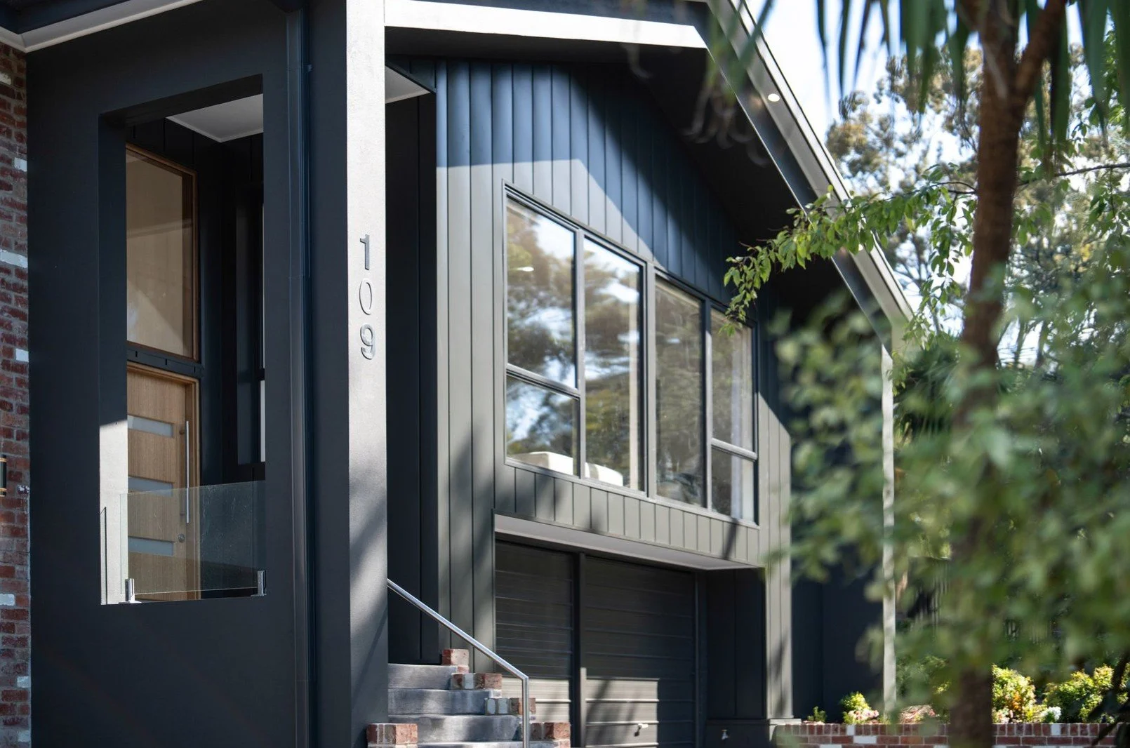 Modern house with dark gray exterior, large windows, and a wooden door, with the house number 109 on a concrete pillar, surrounded by greenery and trees.