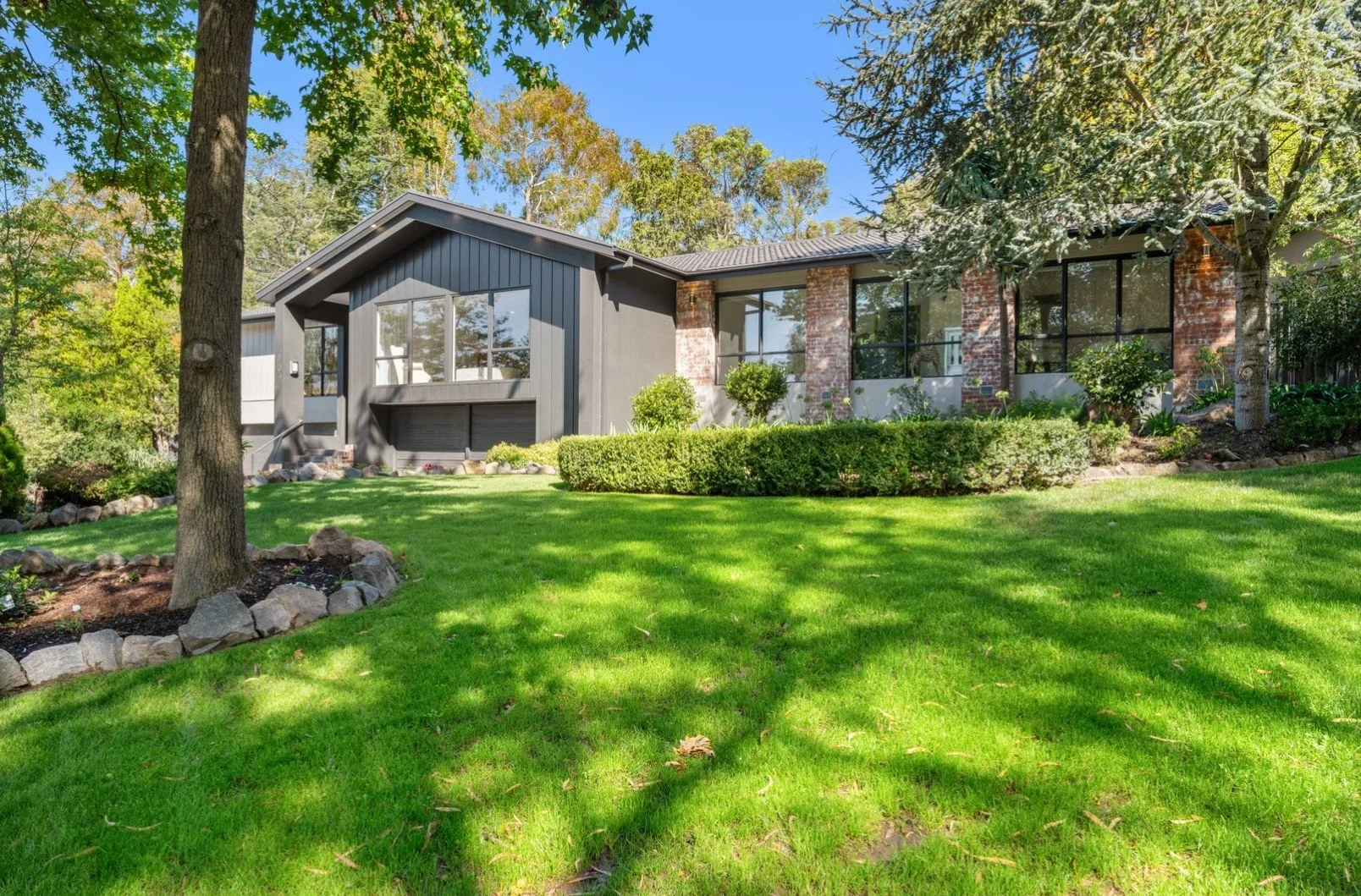 Modern house with large windows, brick accents, and a well-maintained front yard with lush green grass, shrubs, and trees, under a clear blue sky.