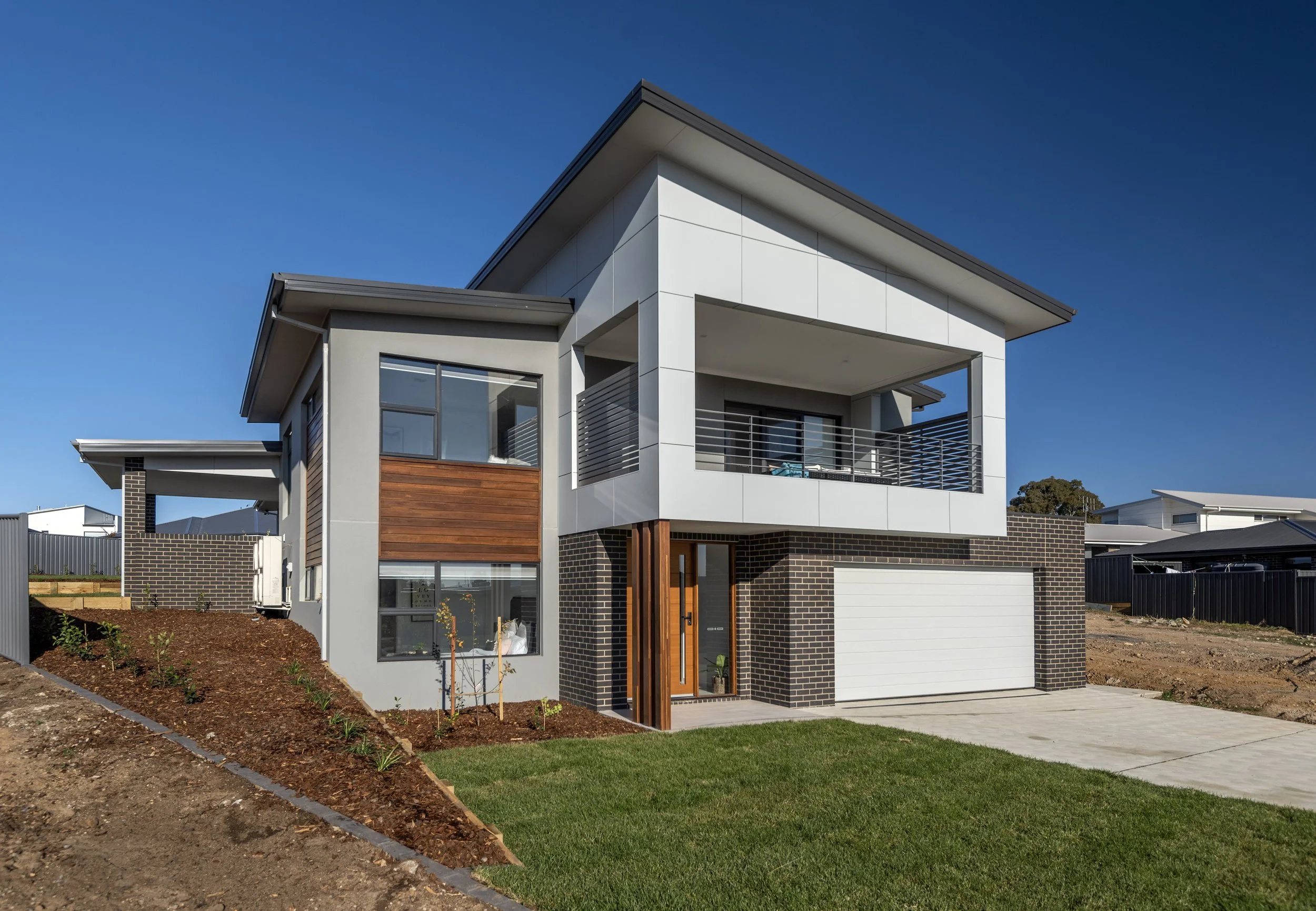 Modern two-story house with white and brick exterior, a balcony, window, and a partly finished front yard with grass and dirt in bright sunlight.