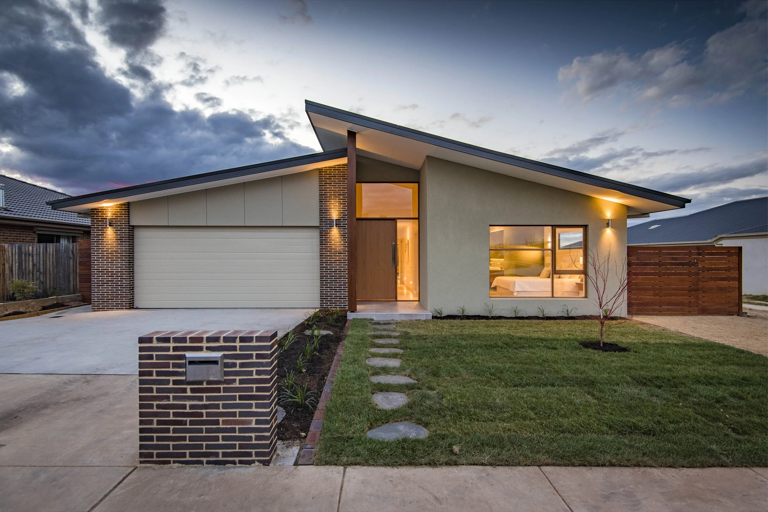 Modern house with brick accents and large windows, illuminated at dusk, front yard with stepping stones and a small tree