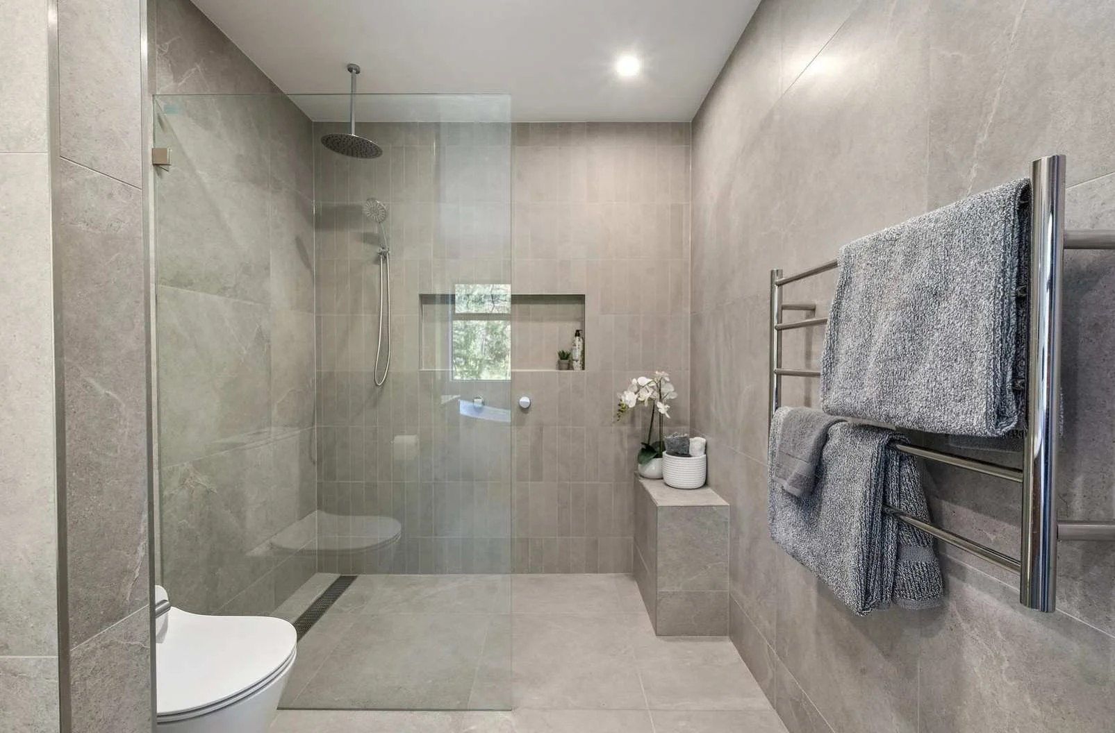 Modern bathroom with walk-in shower, glass enclosure, grey tiled walls and floor, wall-mounted towels rack with grey towels, small ledge with decorative plant and toiletries, and a window with natural light.