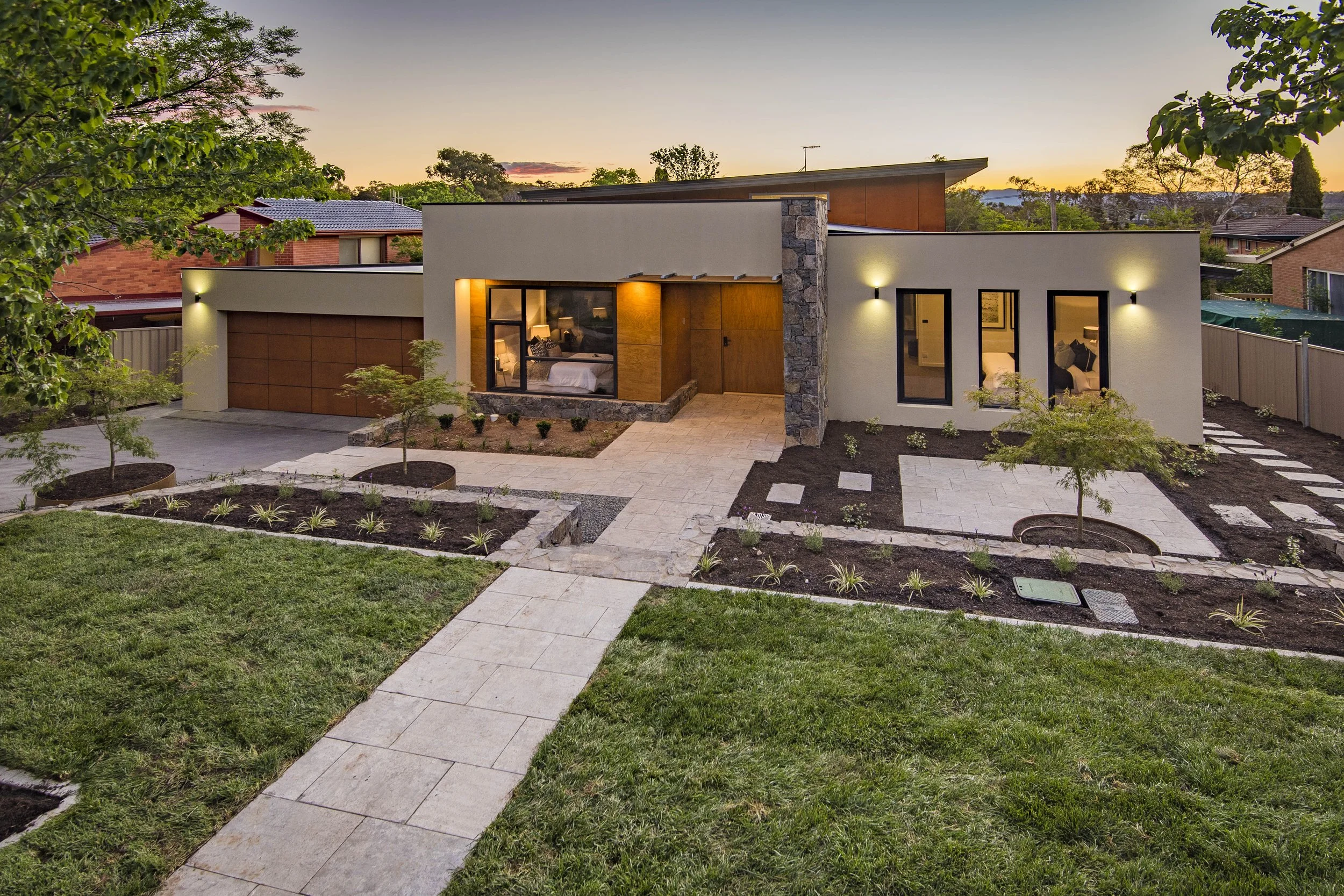 Modern house with a landscaped front yard, stone and wooden accents, large windows, and outdoor lighting at dusk, surrounded by trees and neighboring houses.