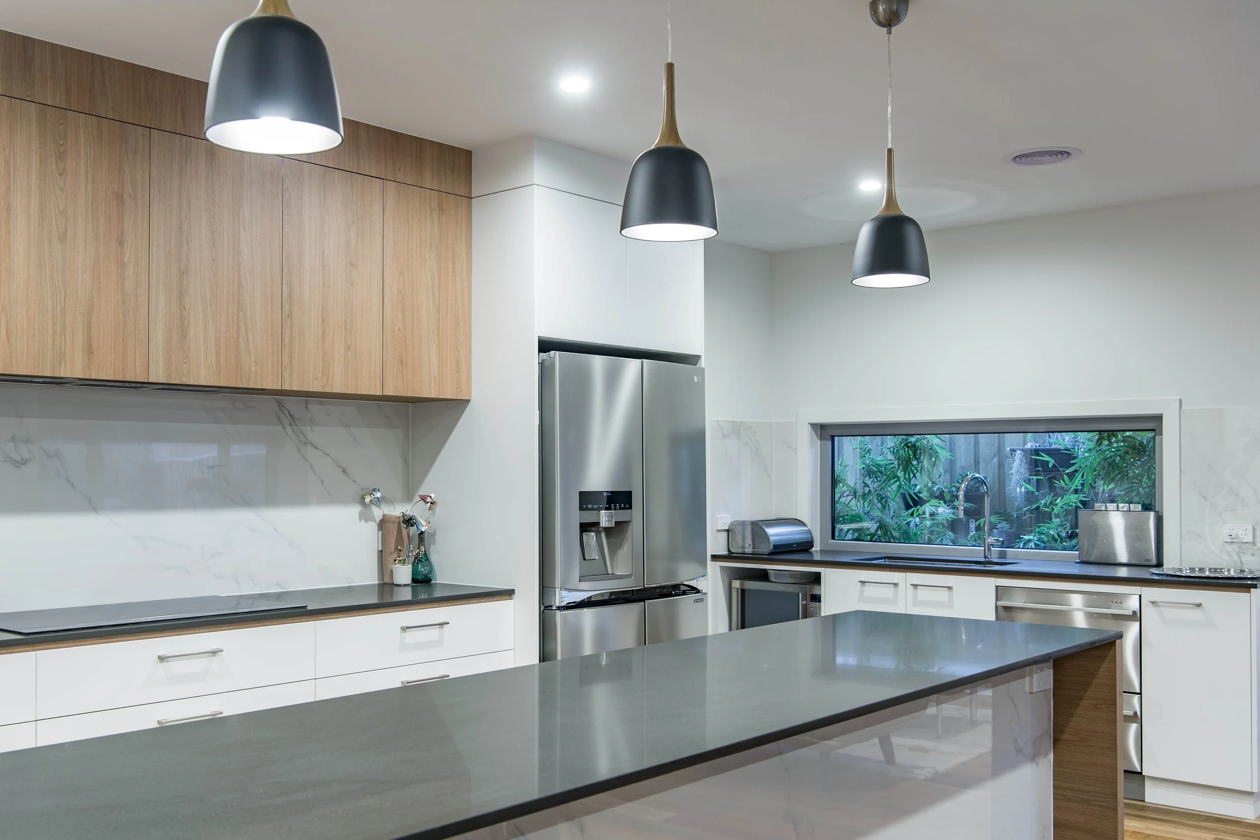 Modern kitchen with white cabinets, wooden upper cabinets, marble backsplash, black countertop, stainless steel refrigerator, window with greenery outside, black pendant lights, and a marble sink.